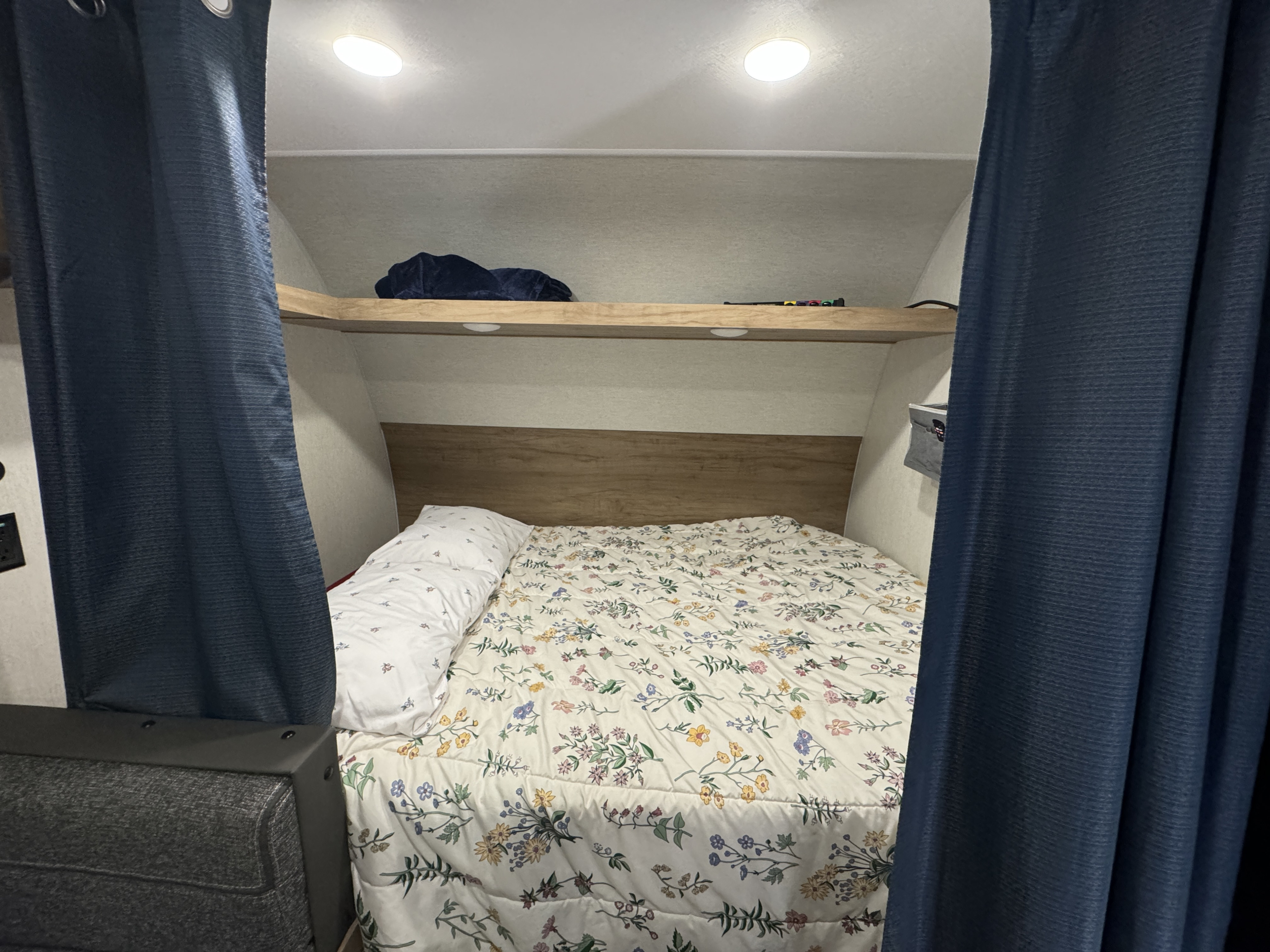 Cozy RV rental bedroom: floral bed, wooden headboard, overhead shelf, and dark blue privacy curtains.. Forest River Cherokee Wolf Den EV16 2024