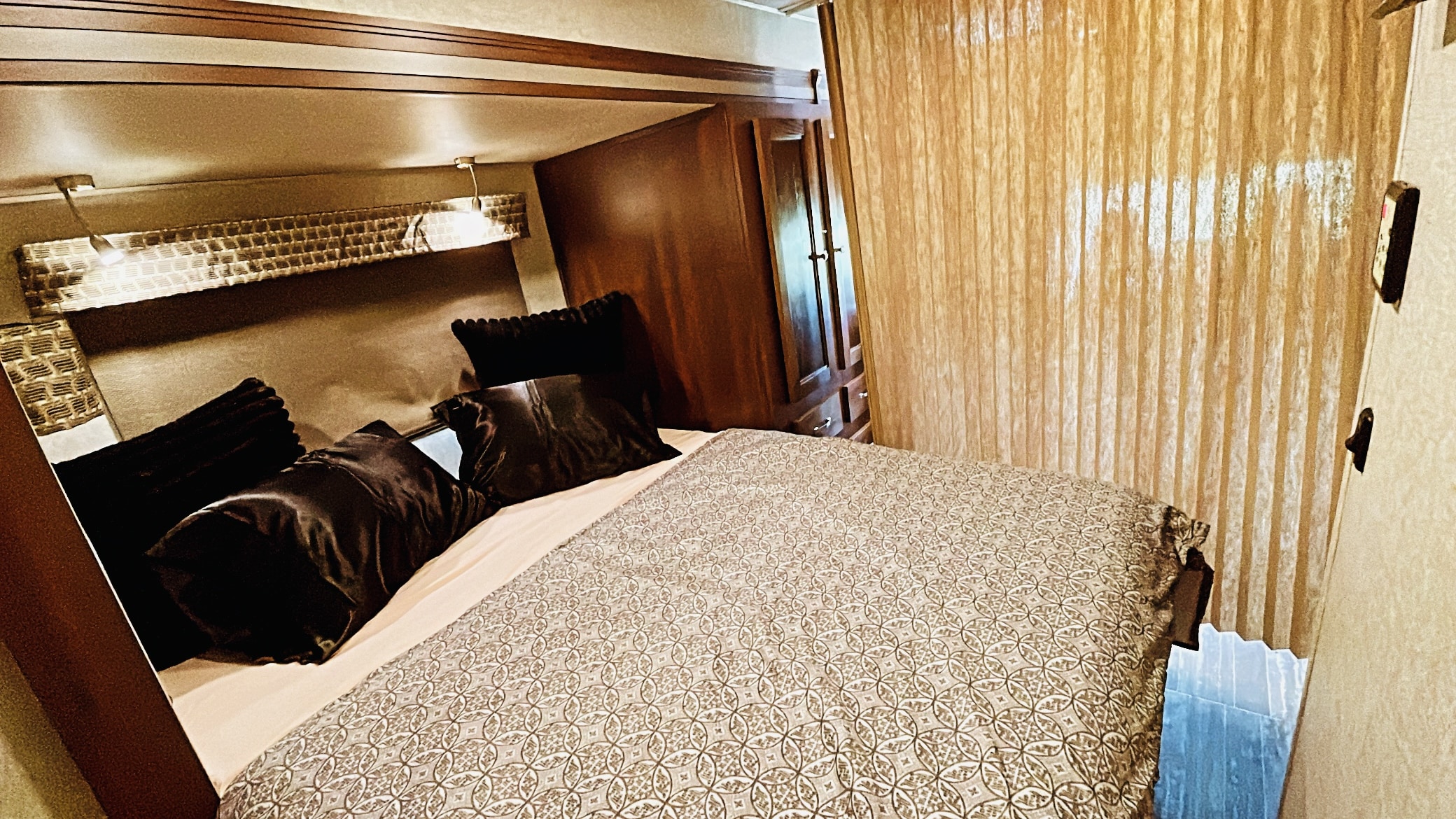 Cozy RV rental bedroom with patterned bed, dark pillows, reading lights, wooden storage, and privacy blinds for your travels.. Forest River Sunseeker MBS 2019