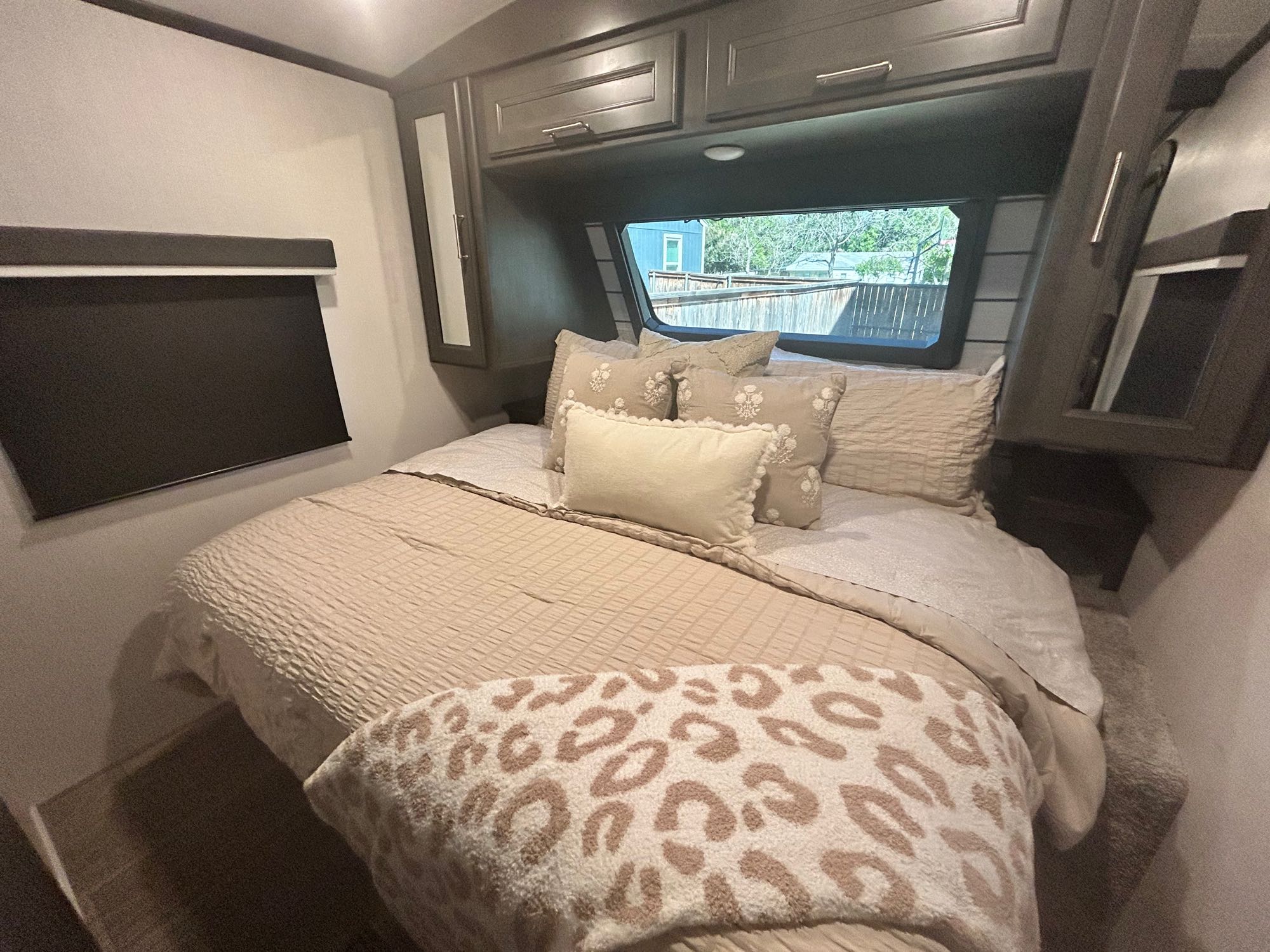 Cozy RV rental bedroom featuring a comfortable bed with light beige bedding, leopard print throw, overhead cabinets, and a window.. Keystone RV Cougar 2024