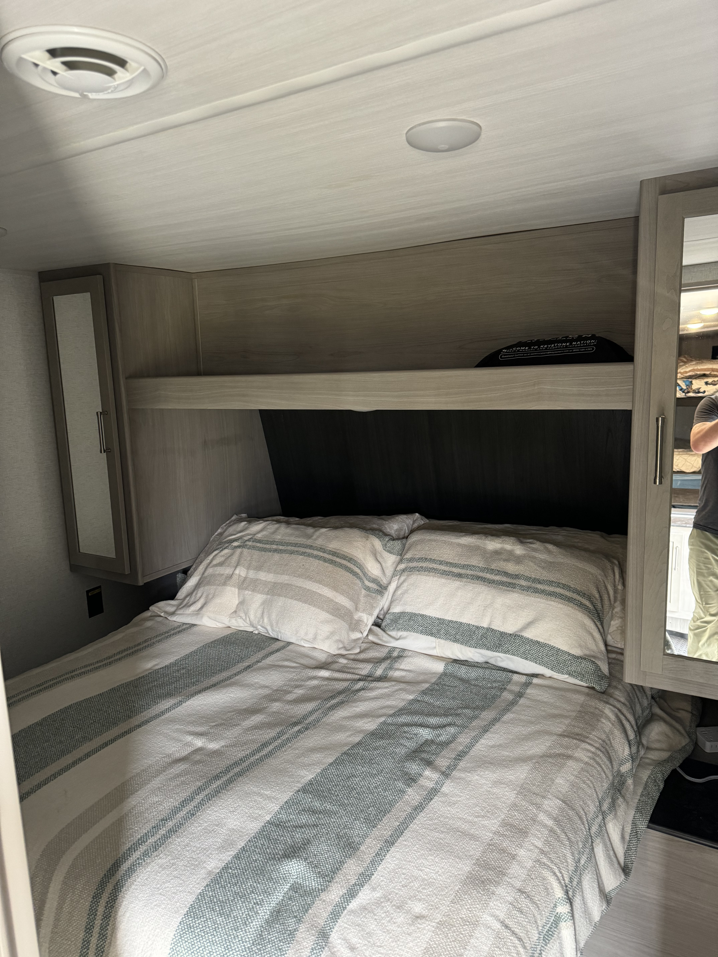 Compact RV rental bedroom featuring a striped bed, built-in storage, and ceiling vent, offering a cozy space.. Keystone RV Springdale 25th Anniversary 2024