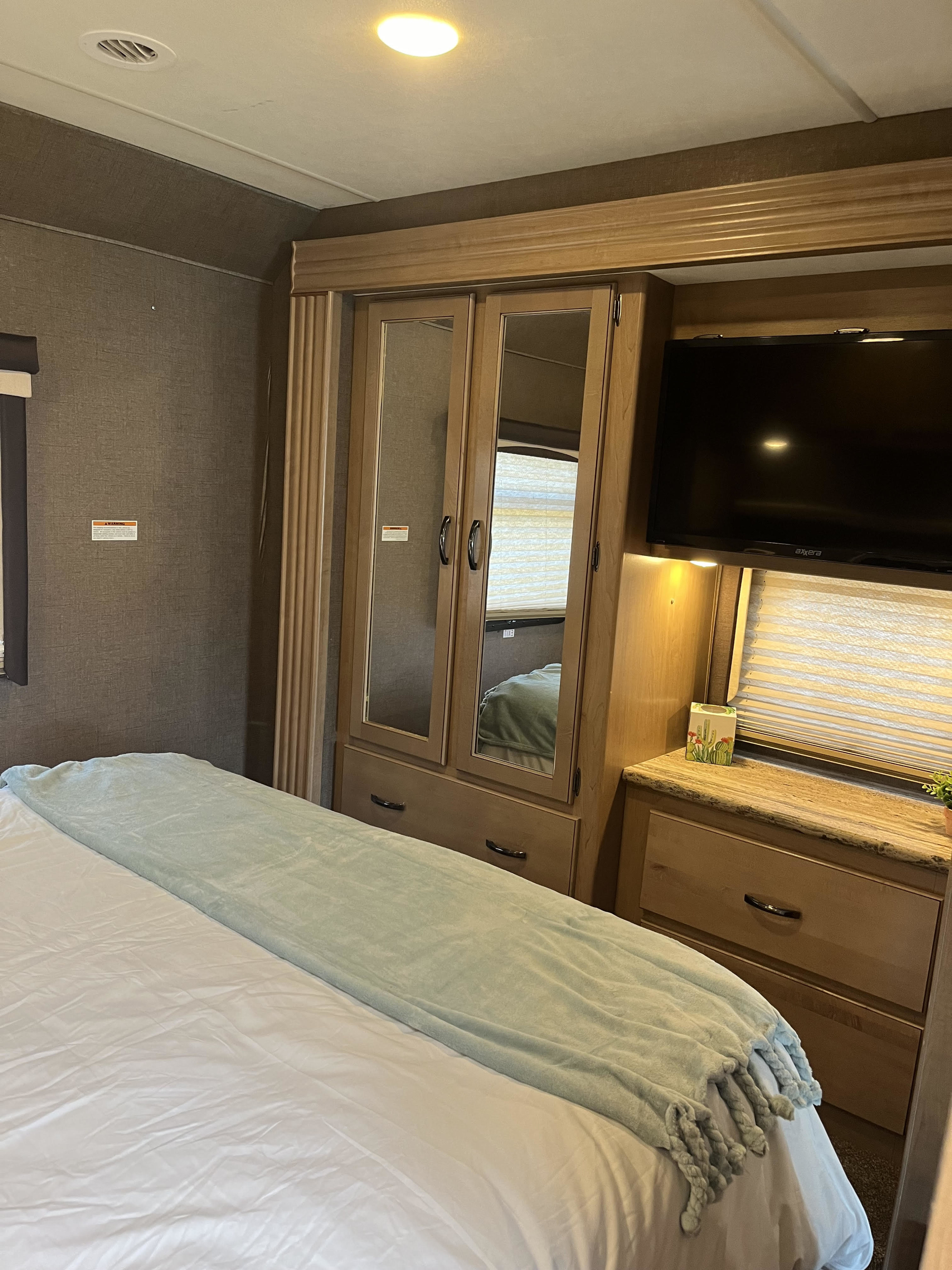 An RV rental bedroom showcasing a bed with white sheets & a light blue blanket, a mirrored wardrobe, TV, and window with blinds.. Thor Motor Coach ACE 2018