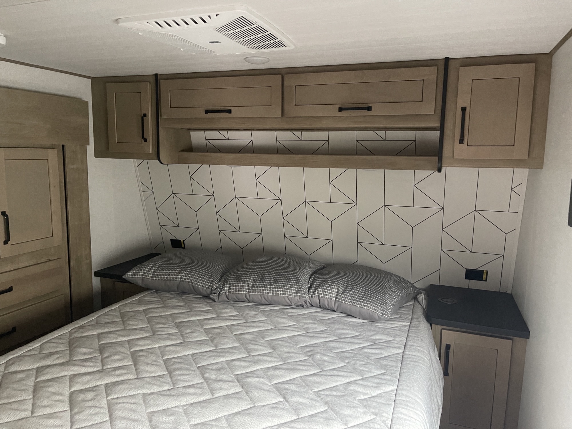 RV rental bedroom: a modern space with geometric headboard, gray bed, wood overhead cabinets, and nightstands.. Keystone Carbon 2022
