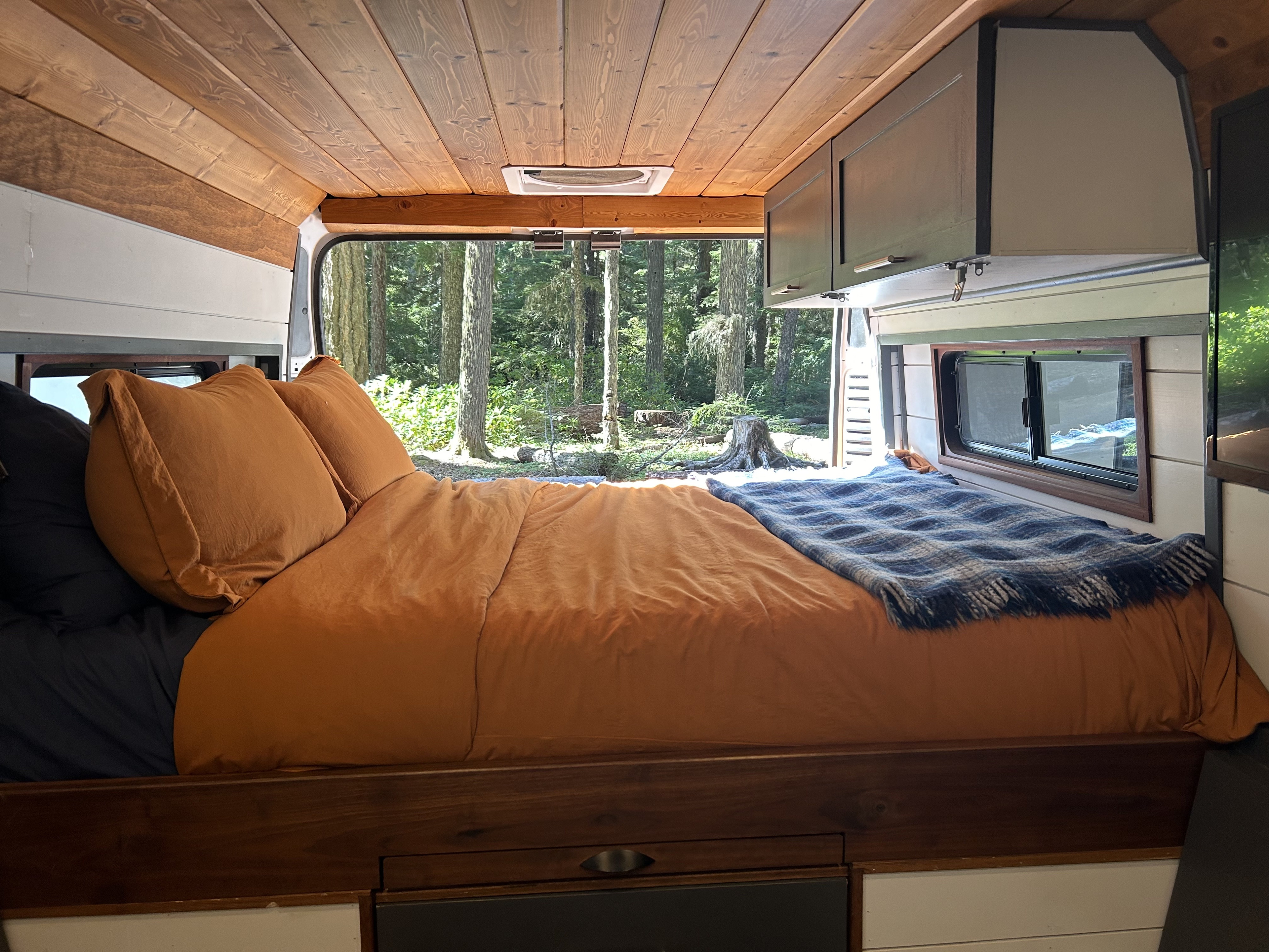 Comfy rv rental interior: bed with orange linens, wooden ceiling, and a serene forest view out the open back doors.. Dodge Ram 3500 2020