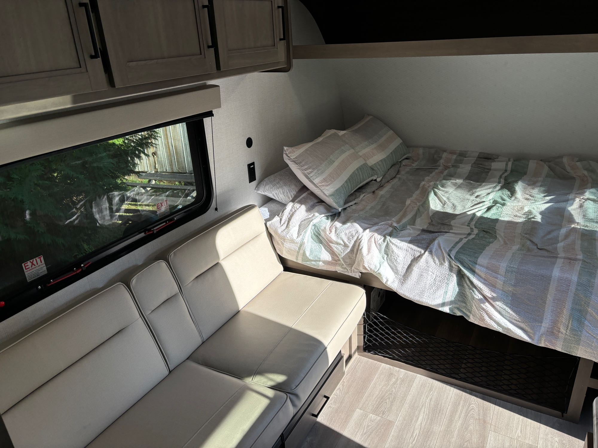 RV rental interior: A bright, cozy space with a neatly made bed featuring striped bedding, a light-colored sofa, and a large window.. Grand Design Transcend One 151BH 2025