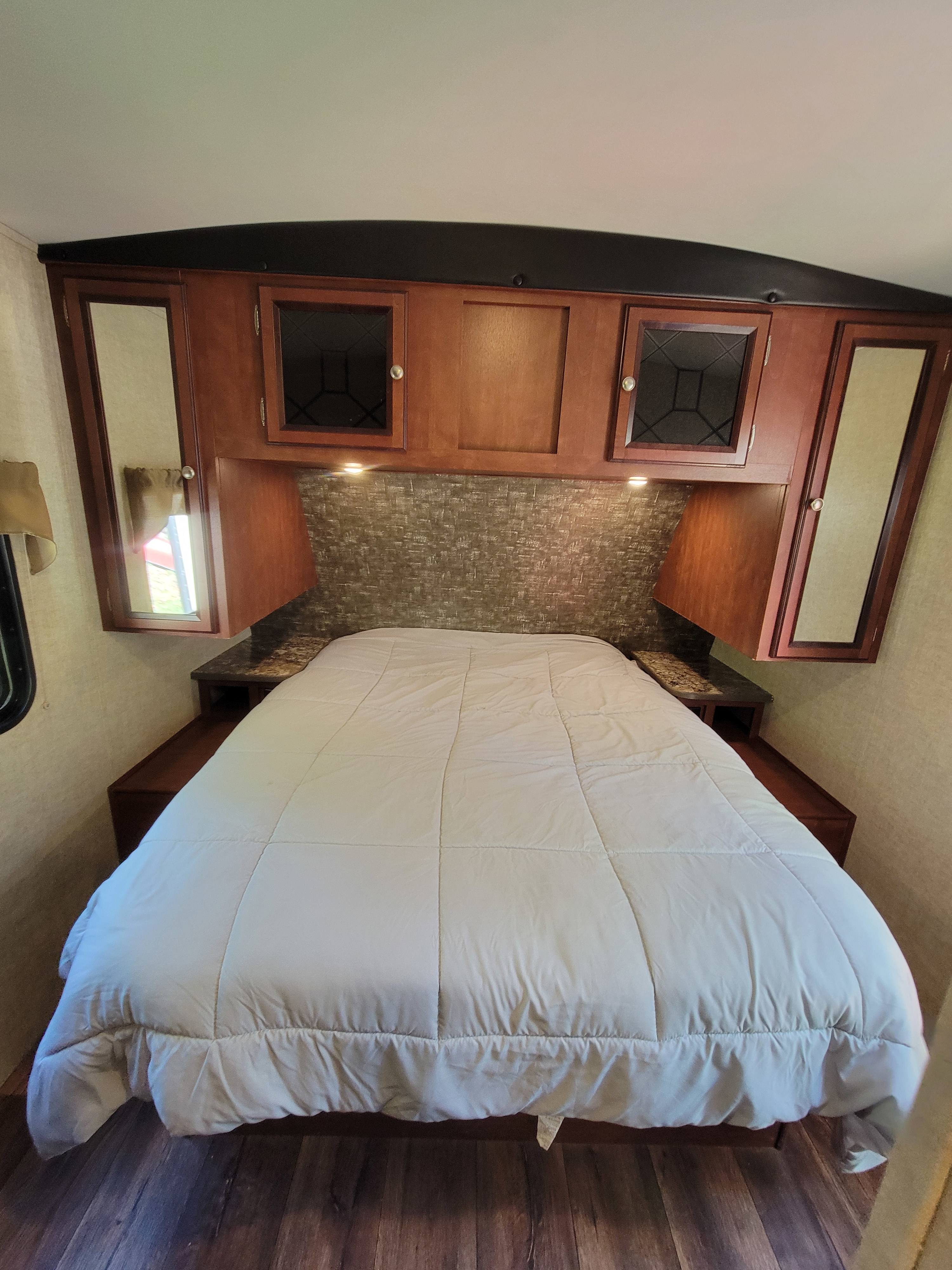 RV rental bedroom: A cozy, full-size bed with overhead and side storage cabinets, a textured headboard, and wood flooring.. Heartland RVs Wilderness 2015