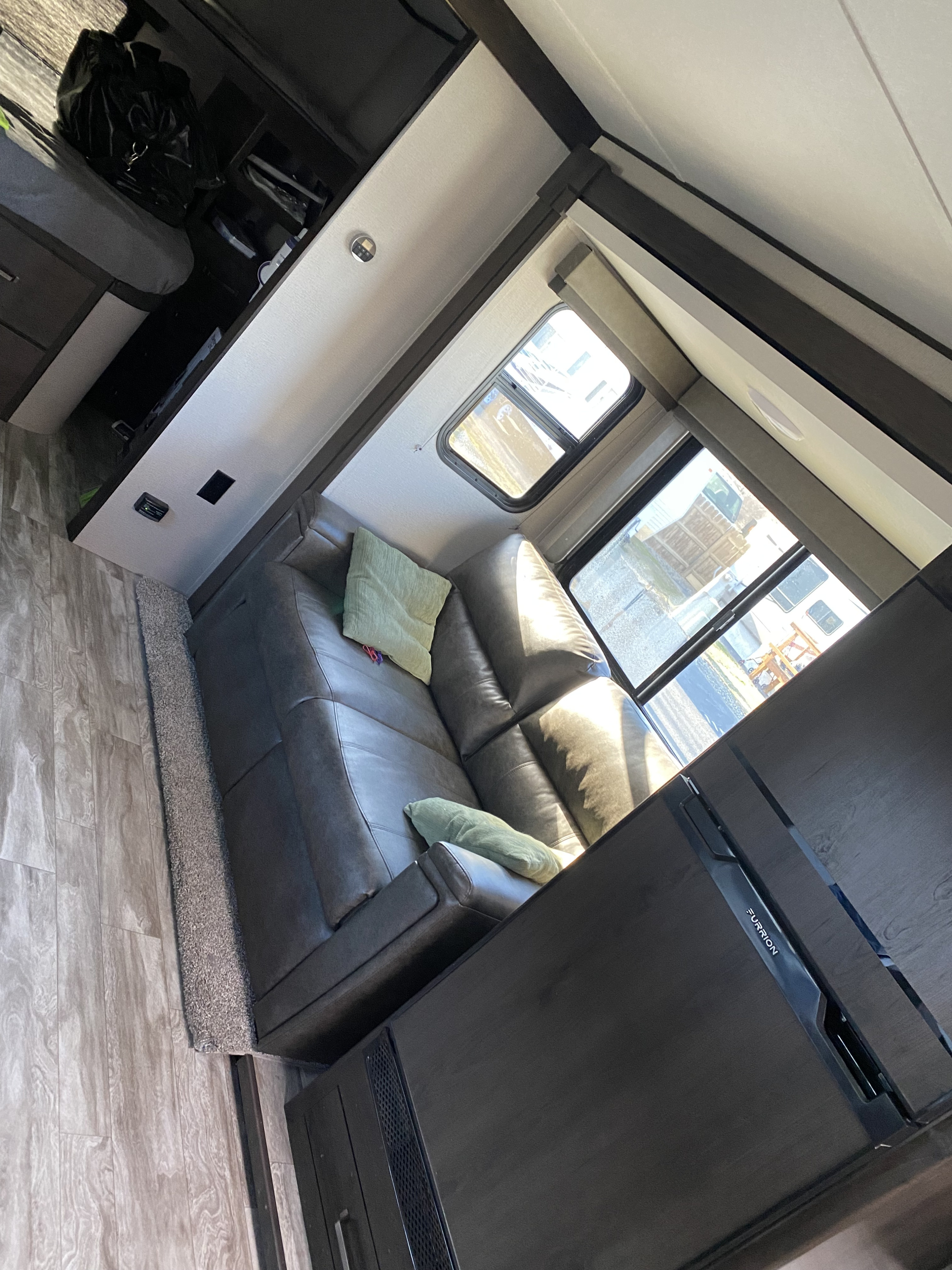 Inside a comfy RV rental: grey sofa with green pillows, large windows, and a Furrion fridge. Wood-look flooring completes the inviting travel space.. Grand Design Transcend Xplor 2021