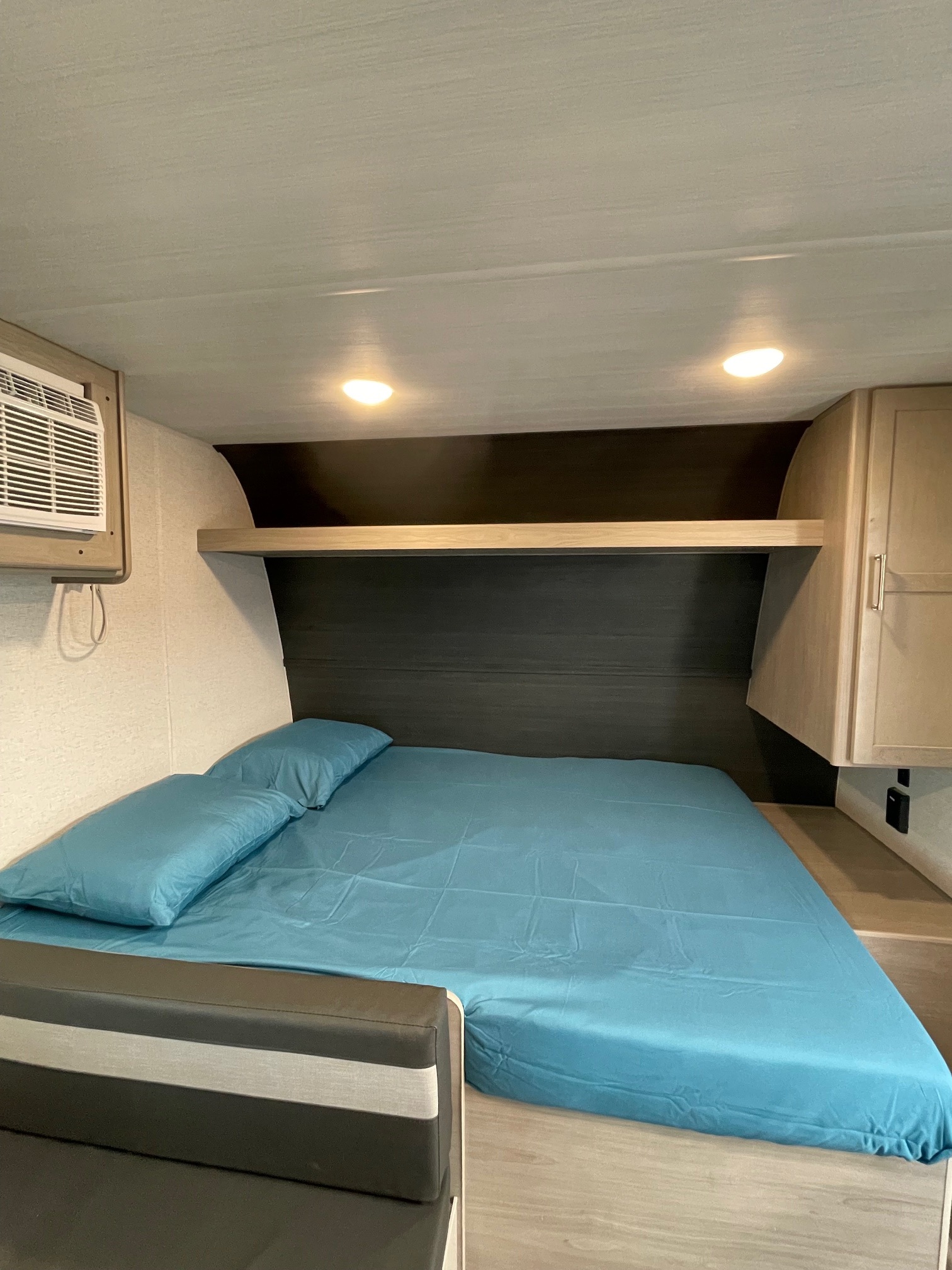 Cozy RV rental bedroom featuring a blue bed with pillows, an overhead shelf, AC unit, and bright lights.. Keystone RV Springdale 2024