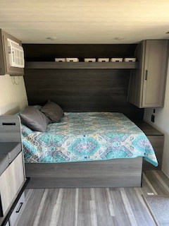 RV rental bedroom: a comfy bed with blue quilt, overhead shelving, side cabinets, and an AC unit on the left.. Keystone RV Hideout 2024