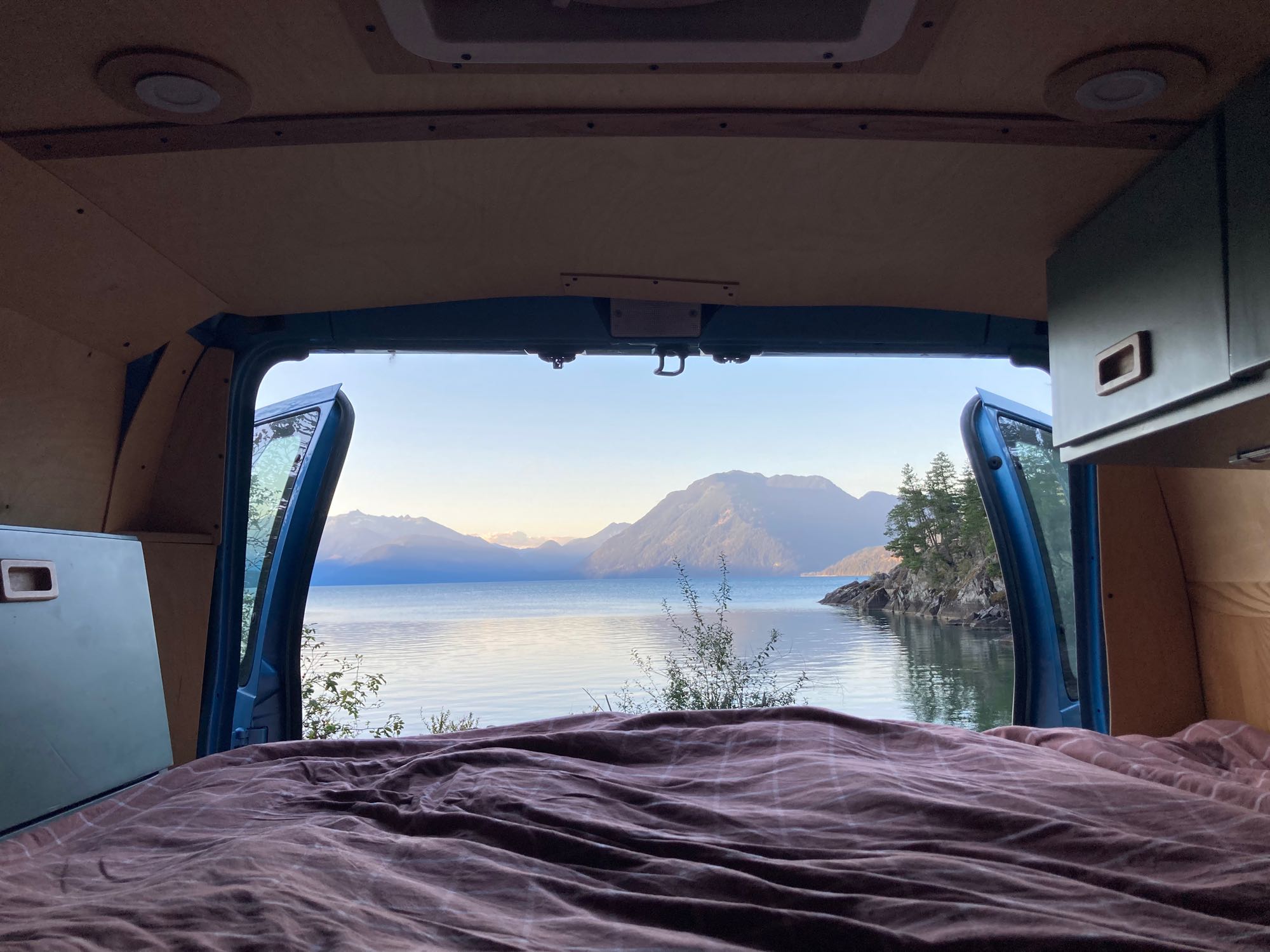 RV rental view: Cozy bed looking out open van doors to a serene lake, mountains, and an island. Perfect van life!. Ford Econoline 2006