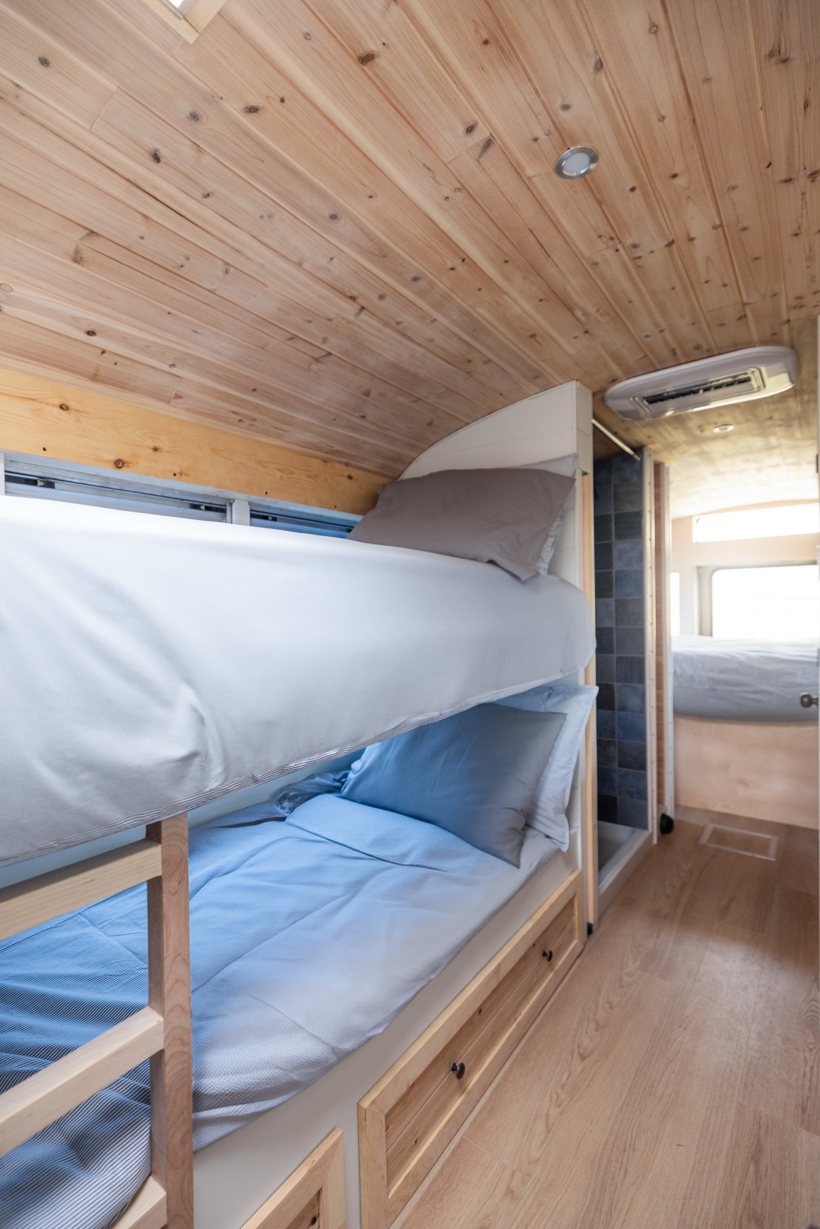 Cozy RV rental interior with wooden bunk beds, built-in storage drawers, and a hallway leading to a second sleeping area.. Inter Skoolie 2007