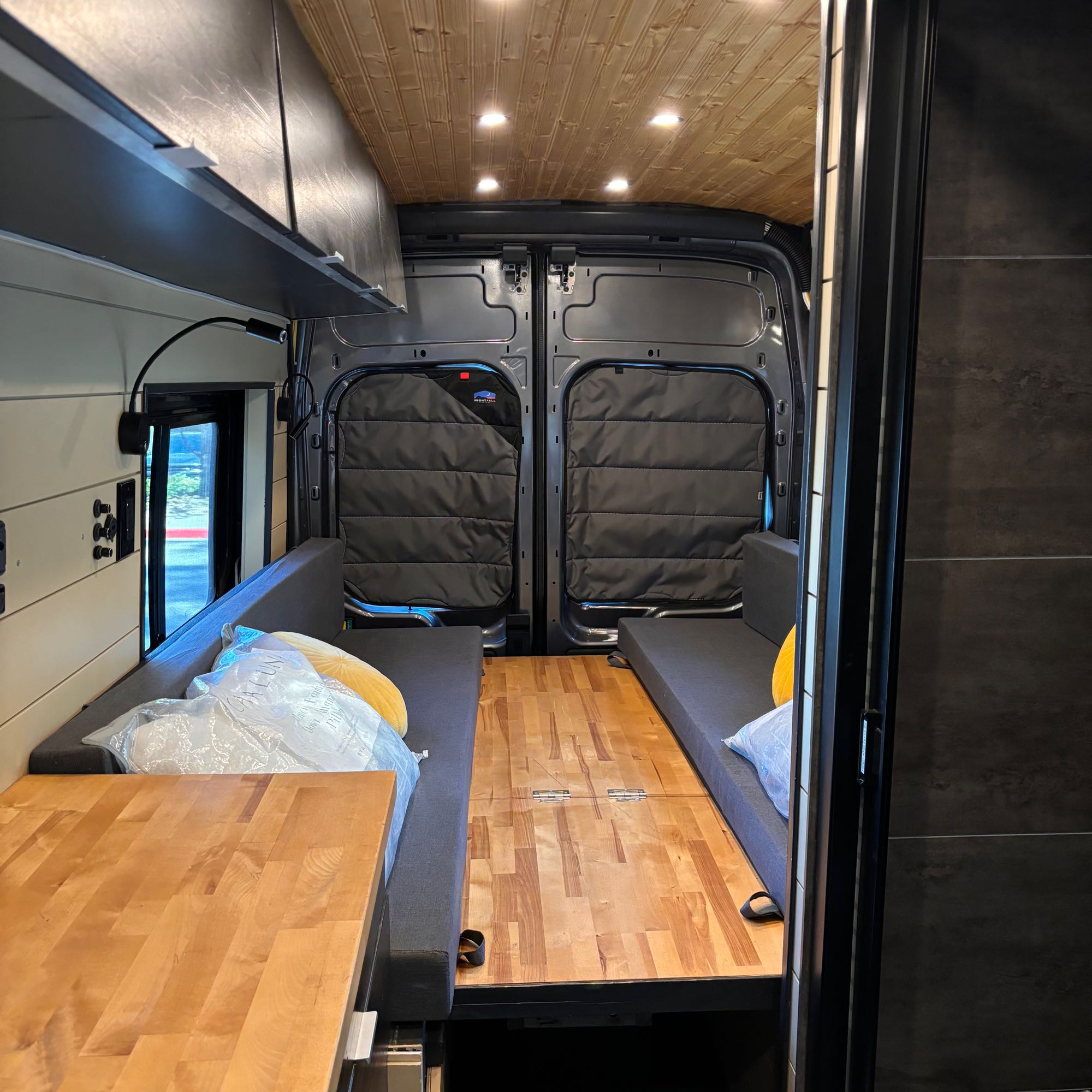 Bright campervan RV rental interior: wood ceiling/floor, grey benches, sleek cabinets, and rear door insulation.. Ford Transit Custom 2023