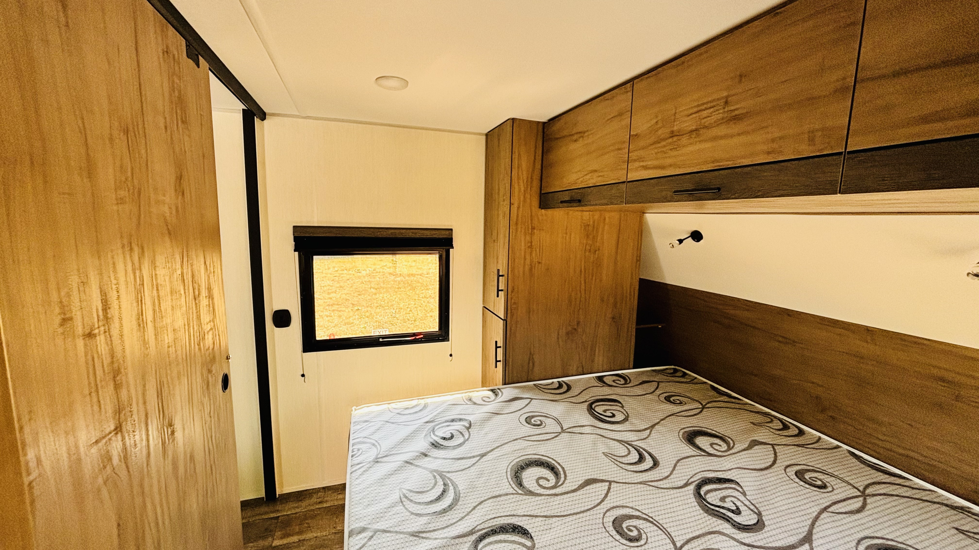 Inside a cozy RV rental bedroom: light walls, wood cabinets, and a patterned bed with a window view.. Forest River Campsite Reserve 27MR 2025