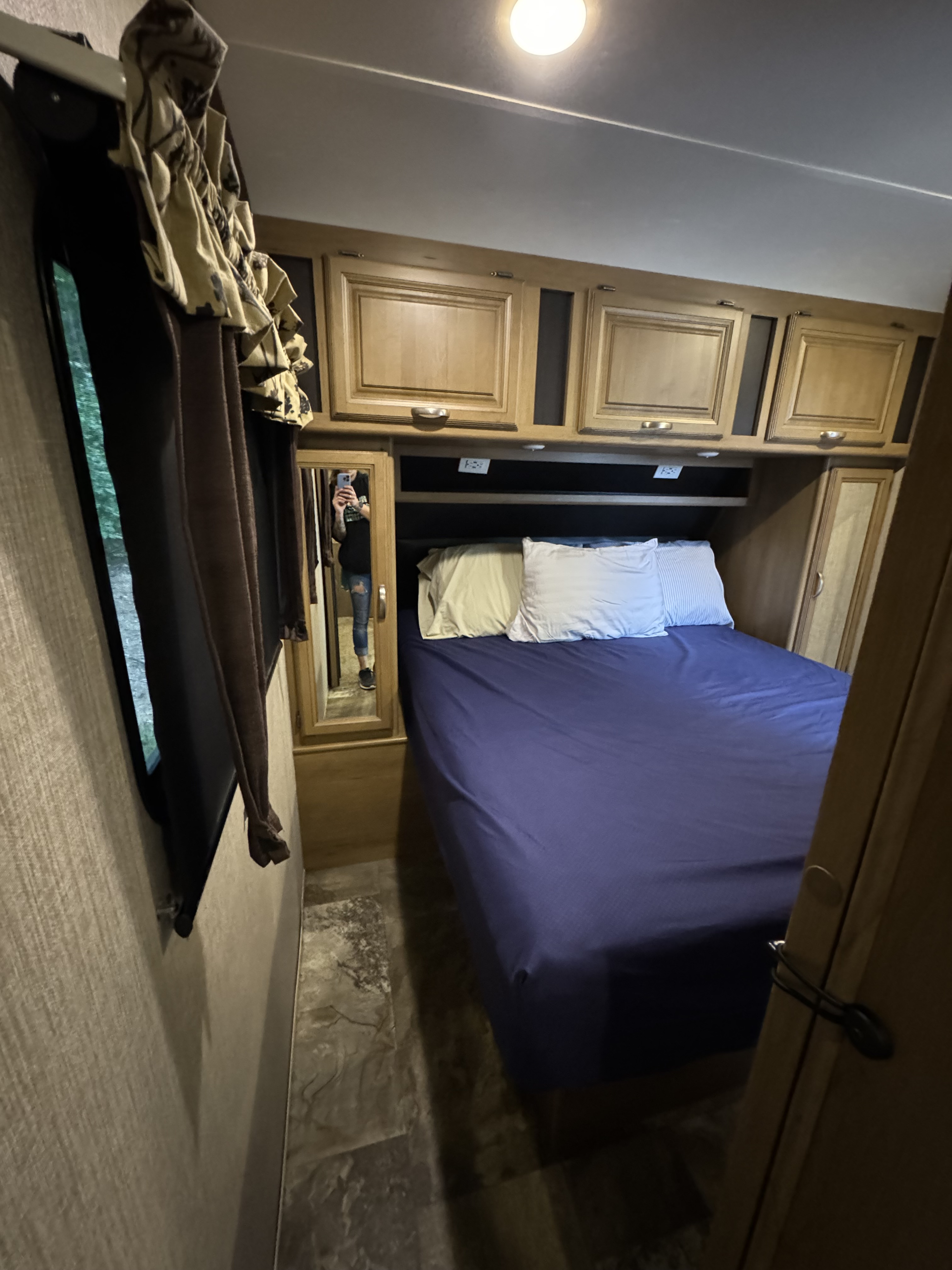 Cozy RV rental bedroom featuring a blue bed, overhead cabinets, window with curtains, and a mirror.. Crossroads RV Cruiser Aire 2016
