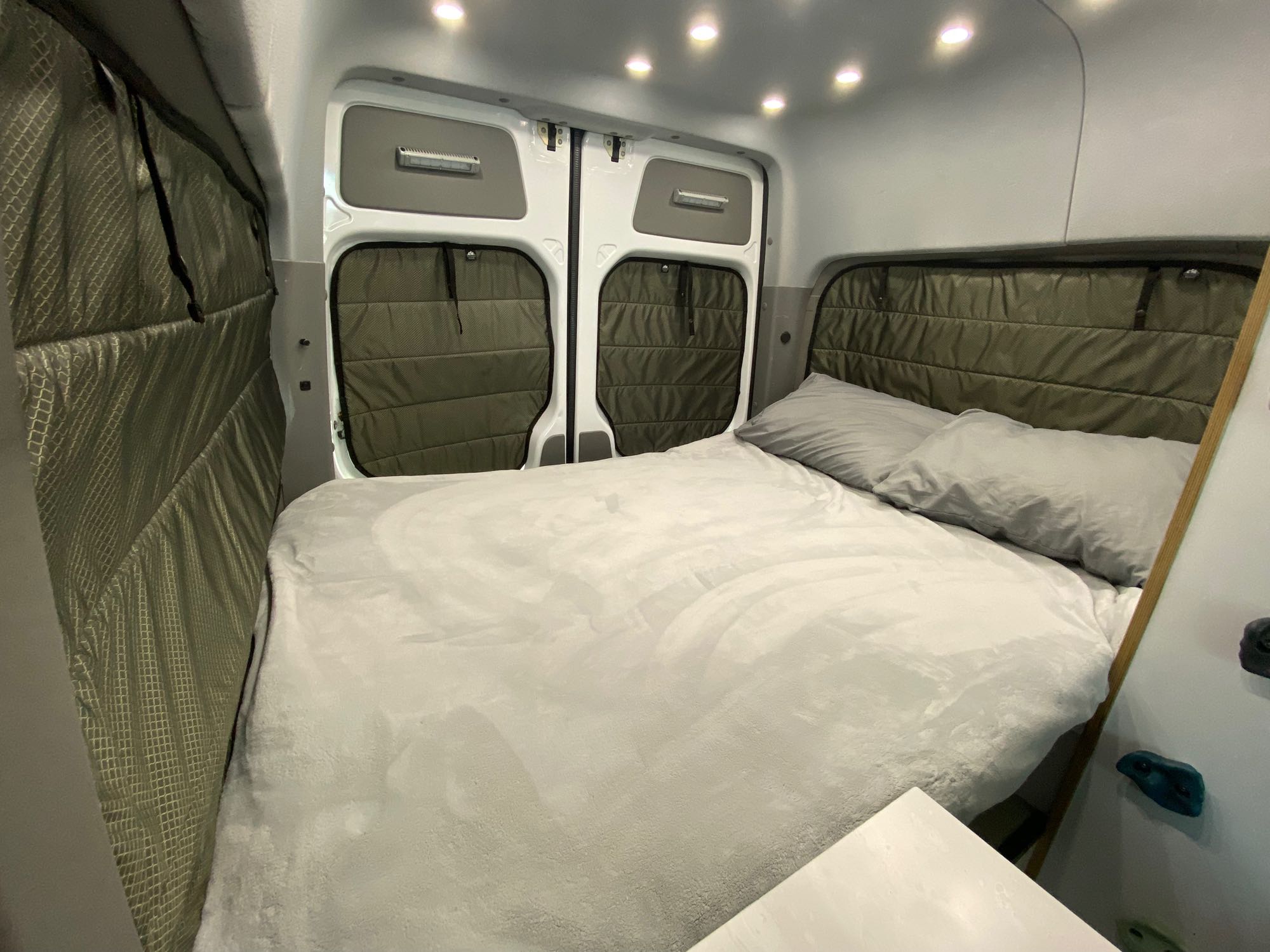 Cozy RV rental bedroom: large bed with light grey bedding, quilted green window insulation, and bright ceiling lights.. Mercedes-Benz Sprinter 2014