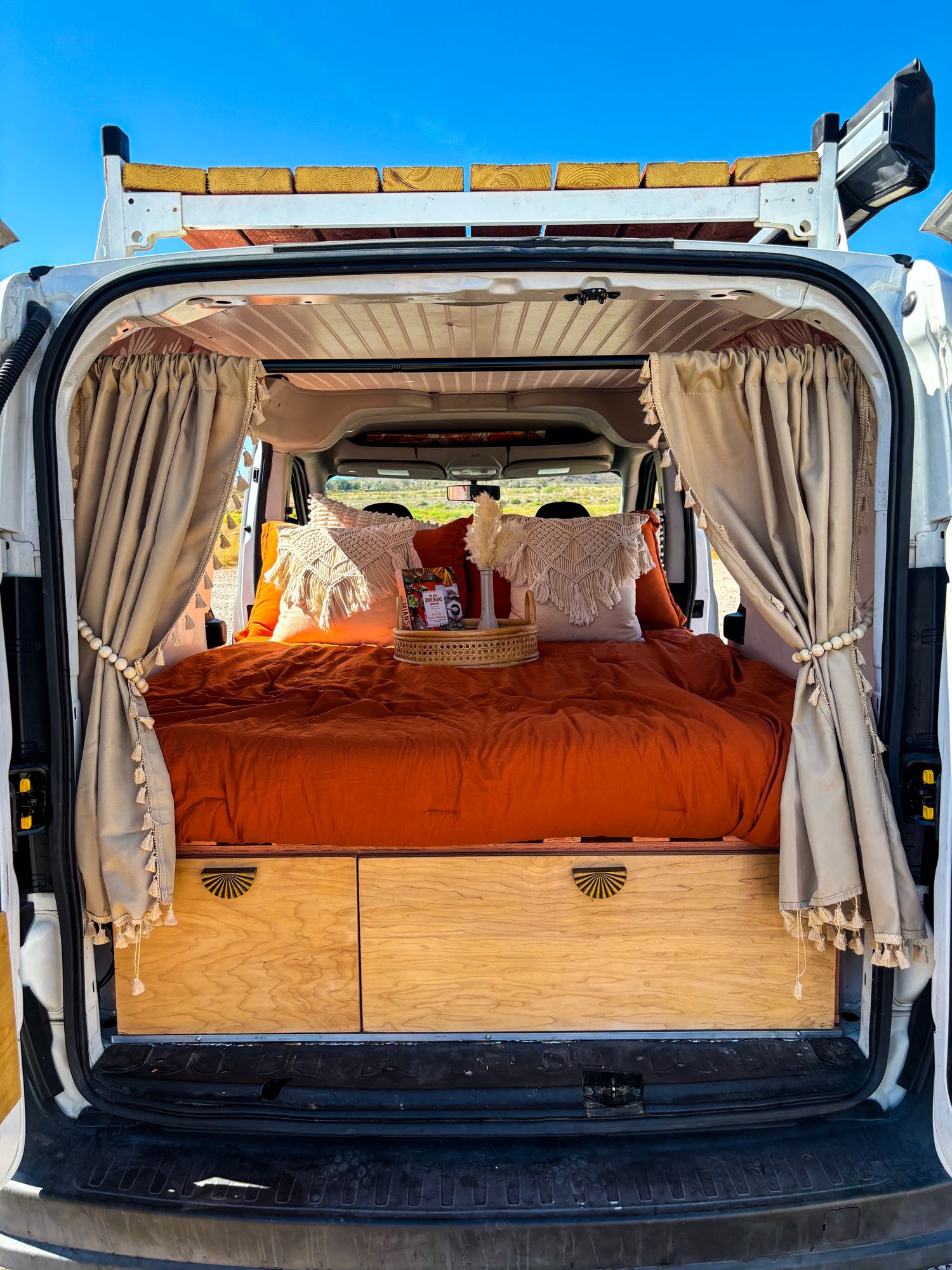 View inside a stylish RV rental van conversion: comfy orange bed, wood storage, and decorative curtains.. Dodge Promaster City 2016