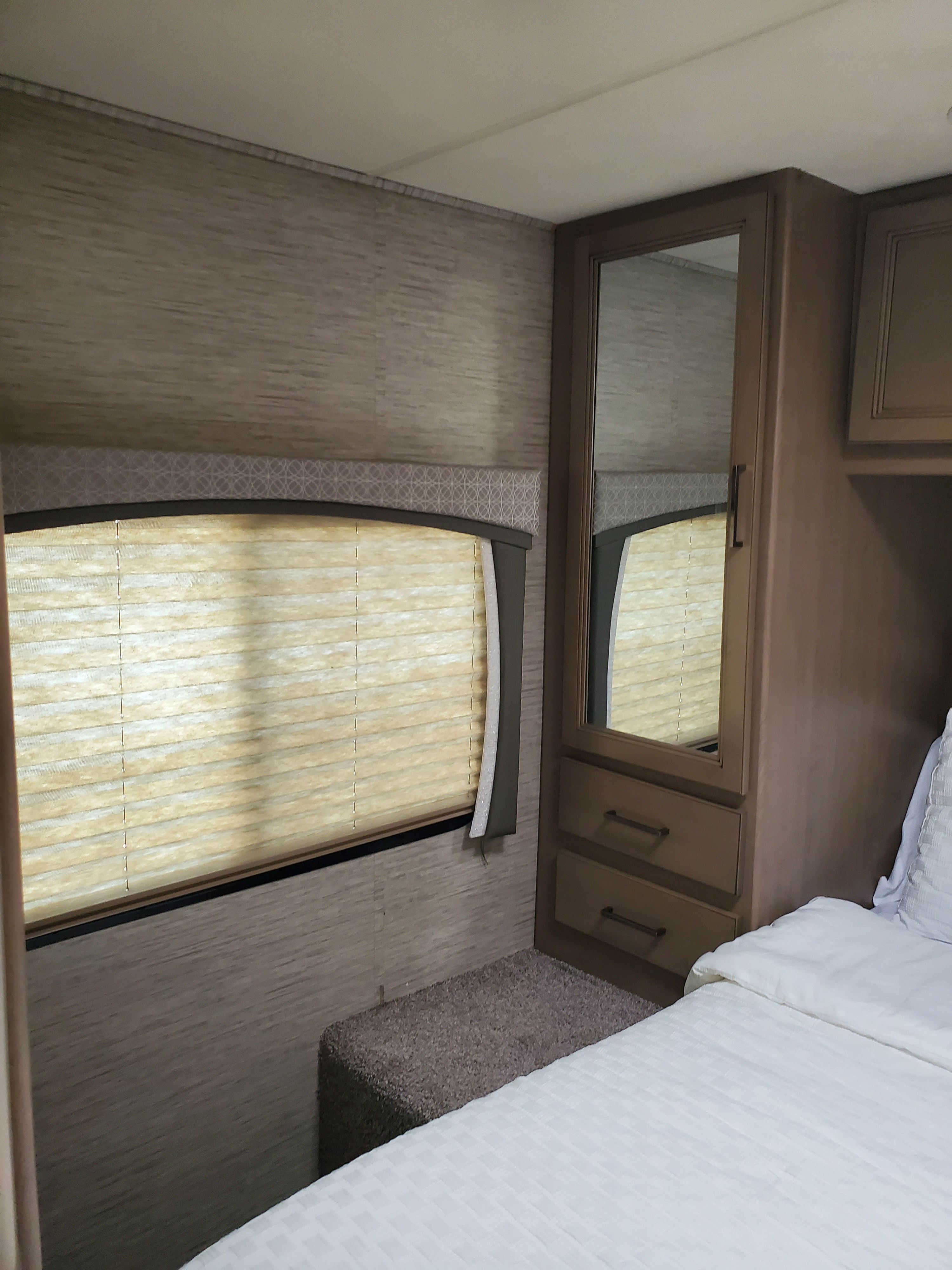 View of an RV rental bedroom: shows a white bed, mirrored wardrobe, and window with light pleated blinds.. Thor Chateau 2021