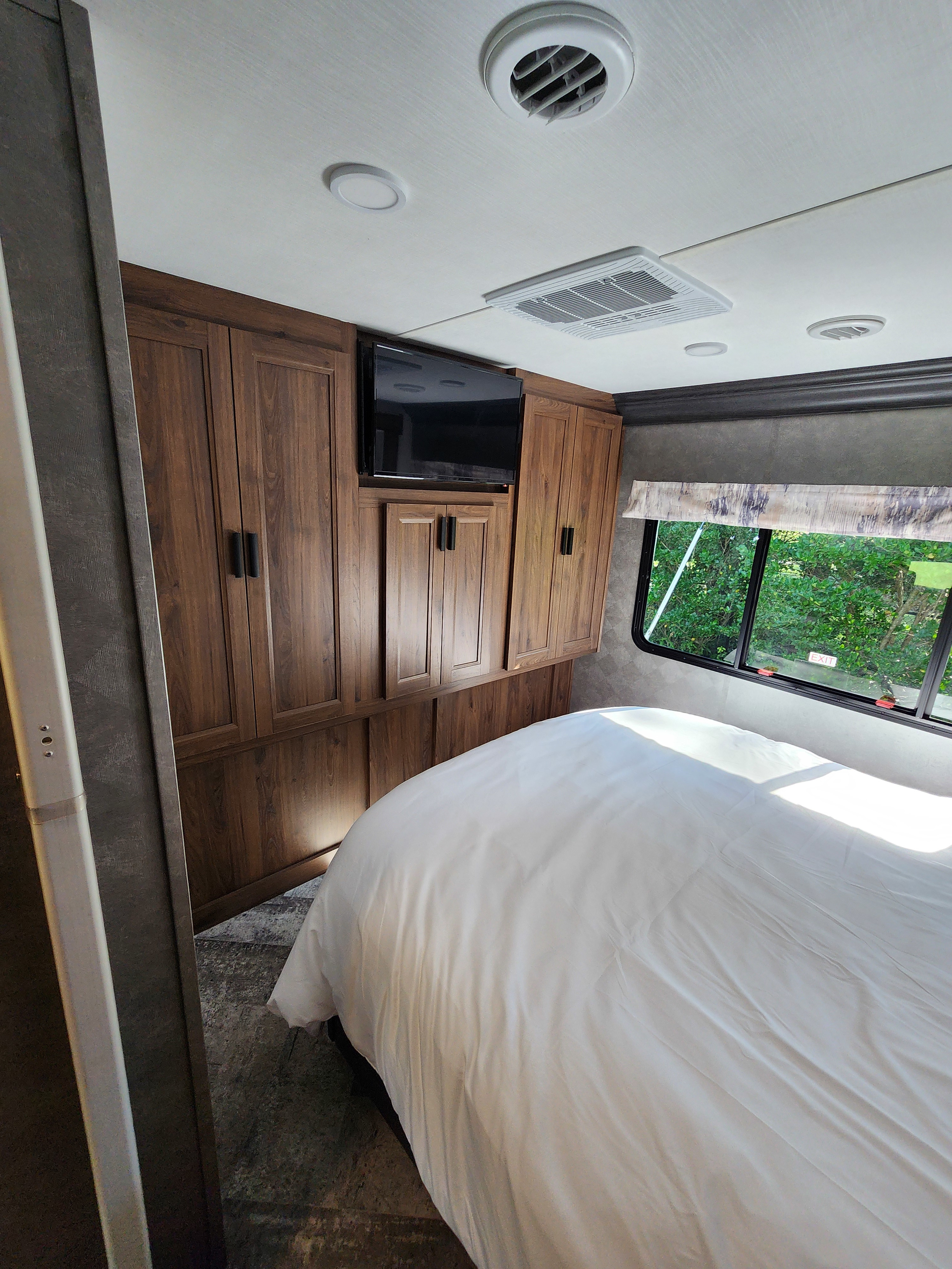 RV rental bedroom: functional space with wood cabinets, TV, AC unit, window, and a crisp white bed. Comfort on the road.. Forest River Sunseeker 2022