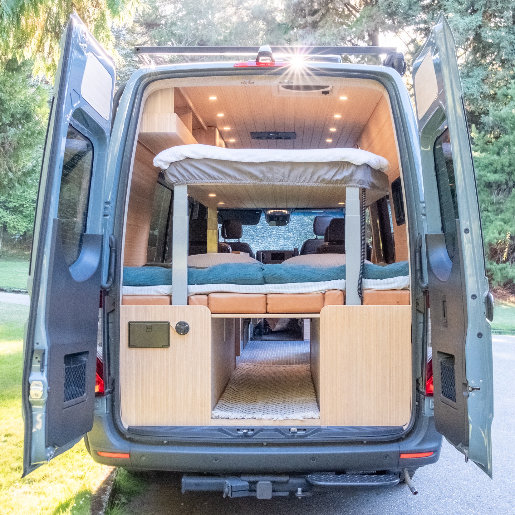 RV rental: Open rear doors of this custom camper van show its cozy interior, featuring elevated & lower beds, and wood paneling.. Mercedes-Benz Sprinter 2021