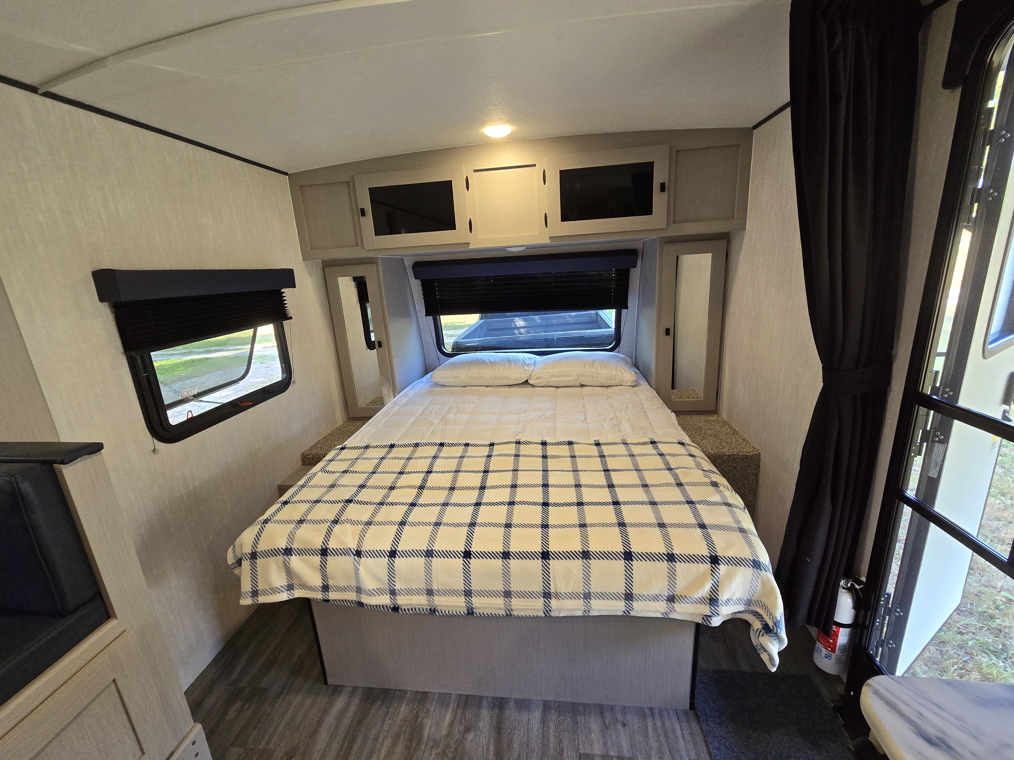 This compact RV rental bedroom features a double bed with a plaid blanket, overhead cabinets, and windows with blinds.. Coachmen Apex Nano 2024