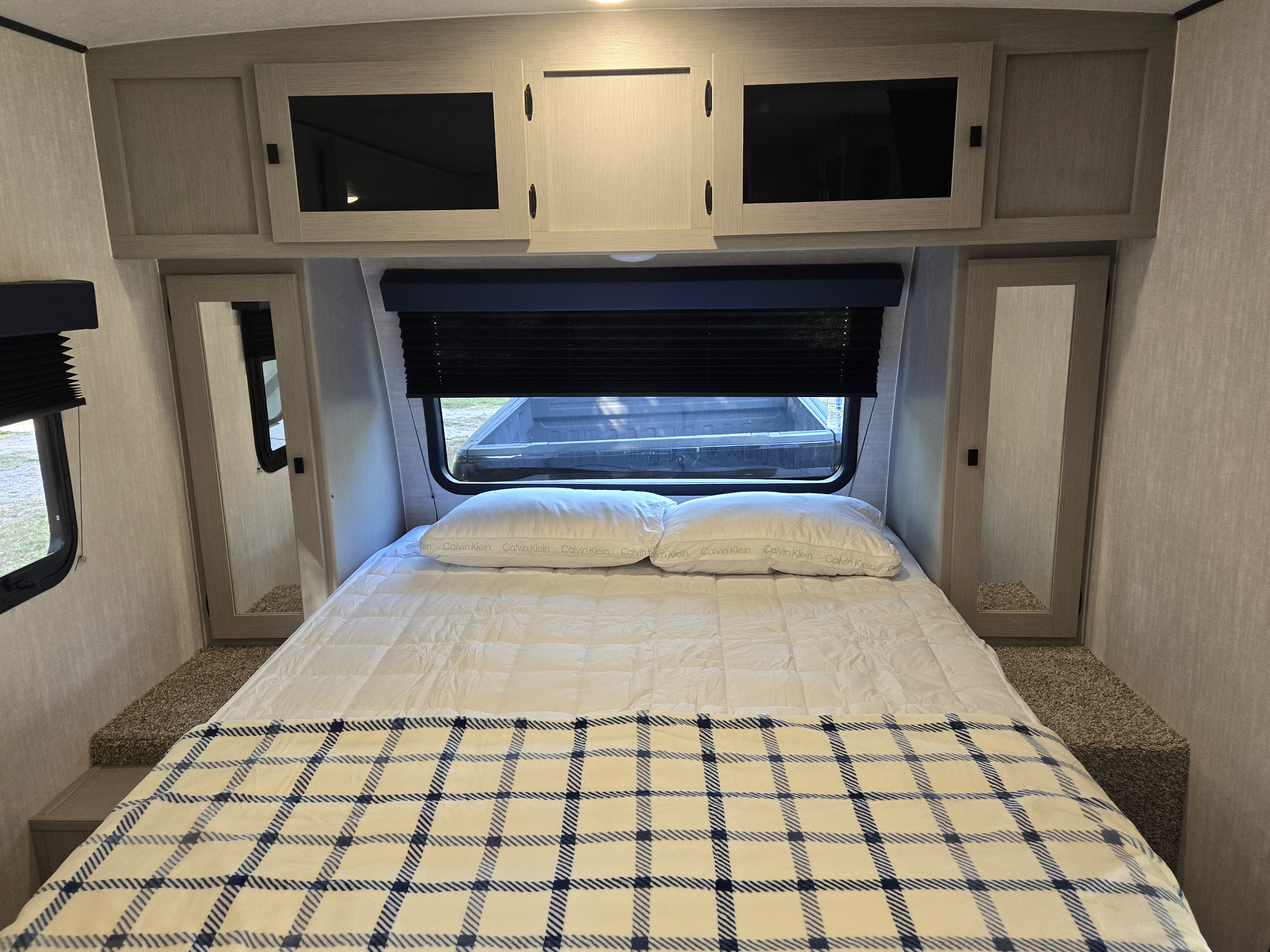 Cozy RV rental bedroom with a queen bed, plaid blanket, overhead cabinets, mirrored wardrobes, and a window with blinds.. Coachmen Apex Nano 2024