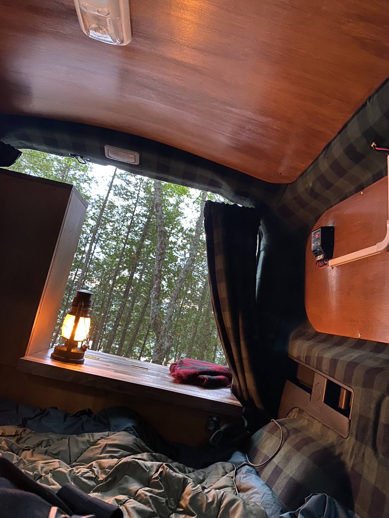 Inside a cozy RV rental: a comfortable bed, a glowing lantern, and a peaceful forest view through the large window.. Dodge Ram Promaster City 2015