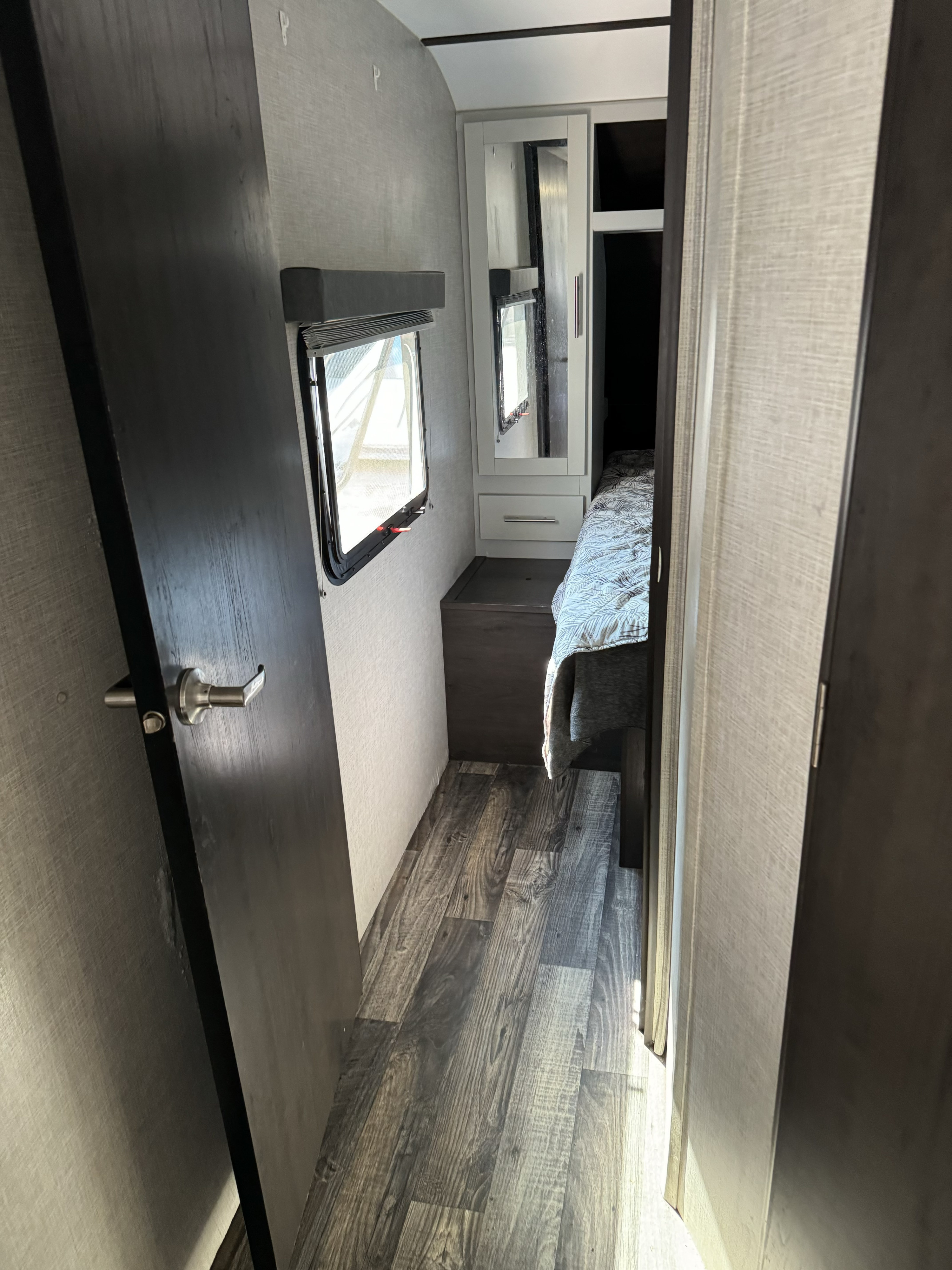 This rv rental features a compact bedroom with a dark door, open window, wood-look floor, and bed by a mirrored cabinet.. Keystone RV Bullet Ultra Lite 2020