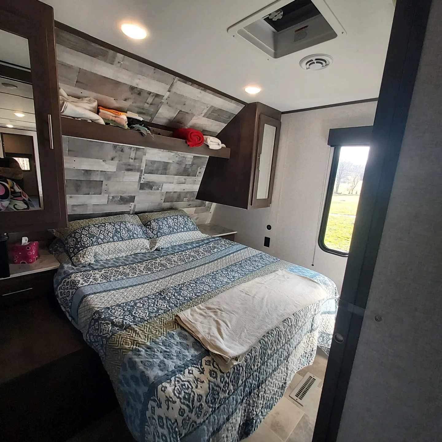 Cozy RV rental bedroom with blue patterned bedding, overhead storage, window view, and light wood accent wall.. Dutchmen Coleman Lantern 2021