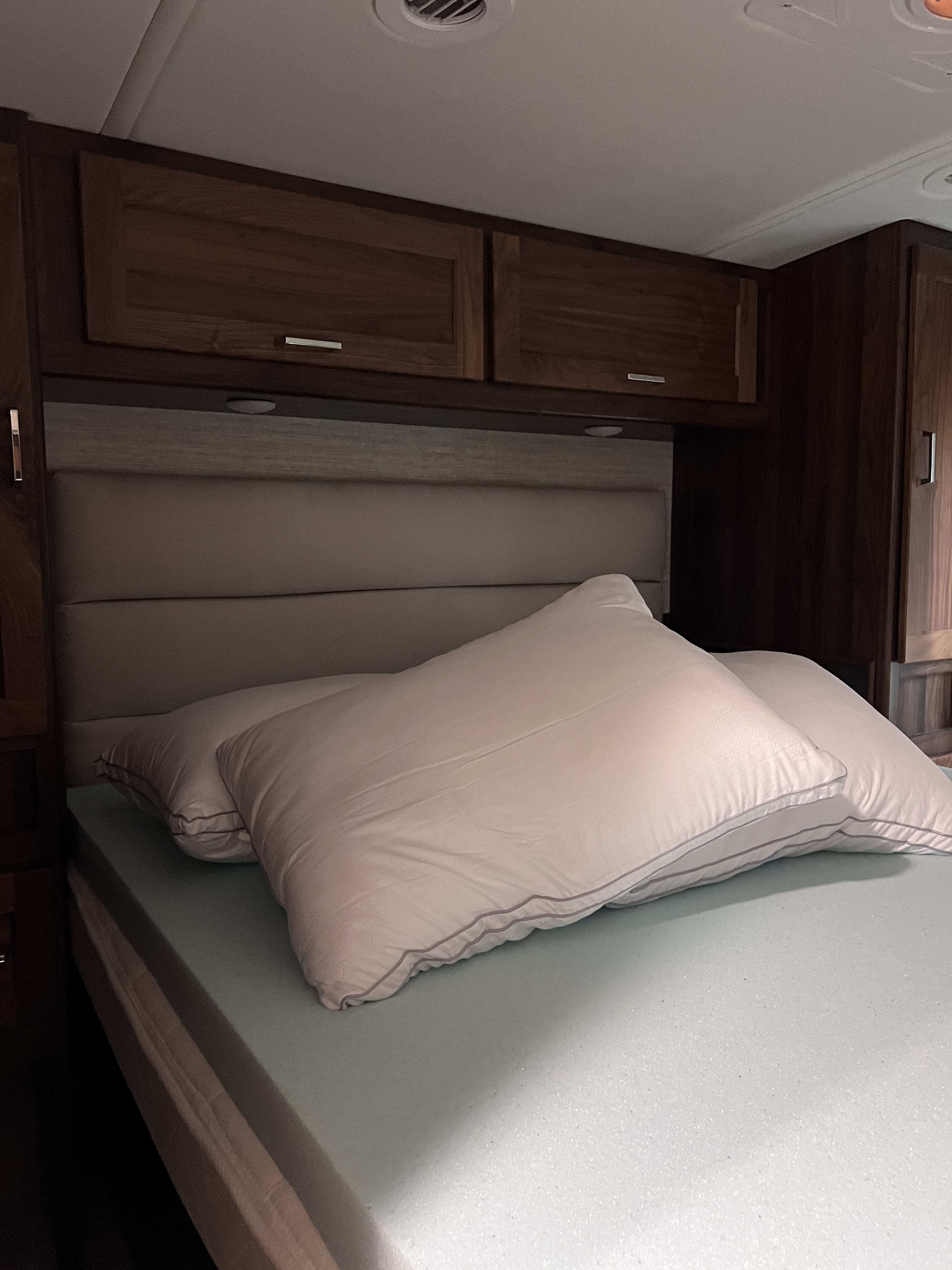 Cozy RV rental bedroom with memory foam mattress, pillows, padded headboard, and wood overhead cabinets for comfortable travel.. Winnebago Vista 2021