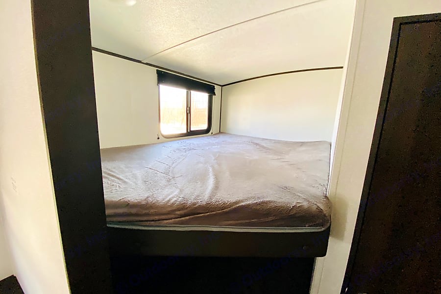 Inside an RV rental, a compact bedroom features a made bed below a window. White walls frame the cozy space.. Keystone Springdale 2021