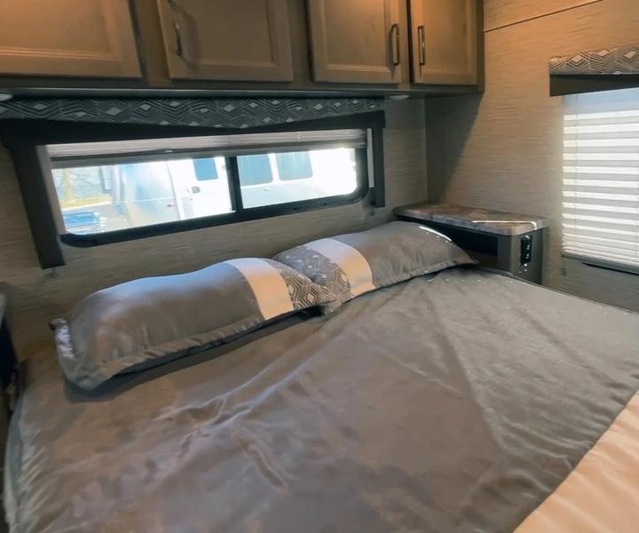 Cozy rv rental bedroom interior featuring a comfortable bed with gray bedding, overhead cabinets, and a window view.. Thor Four Winds 2023