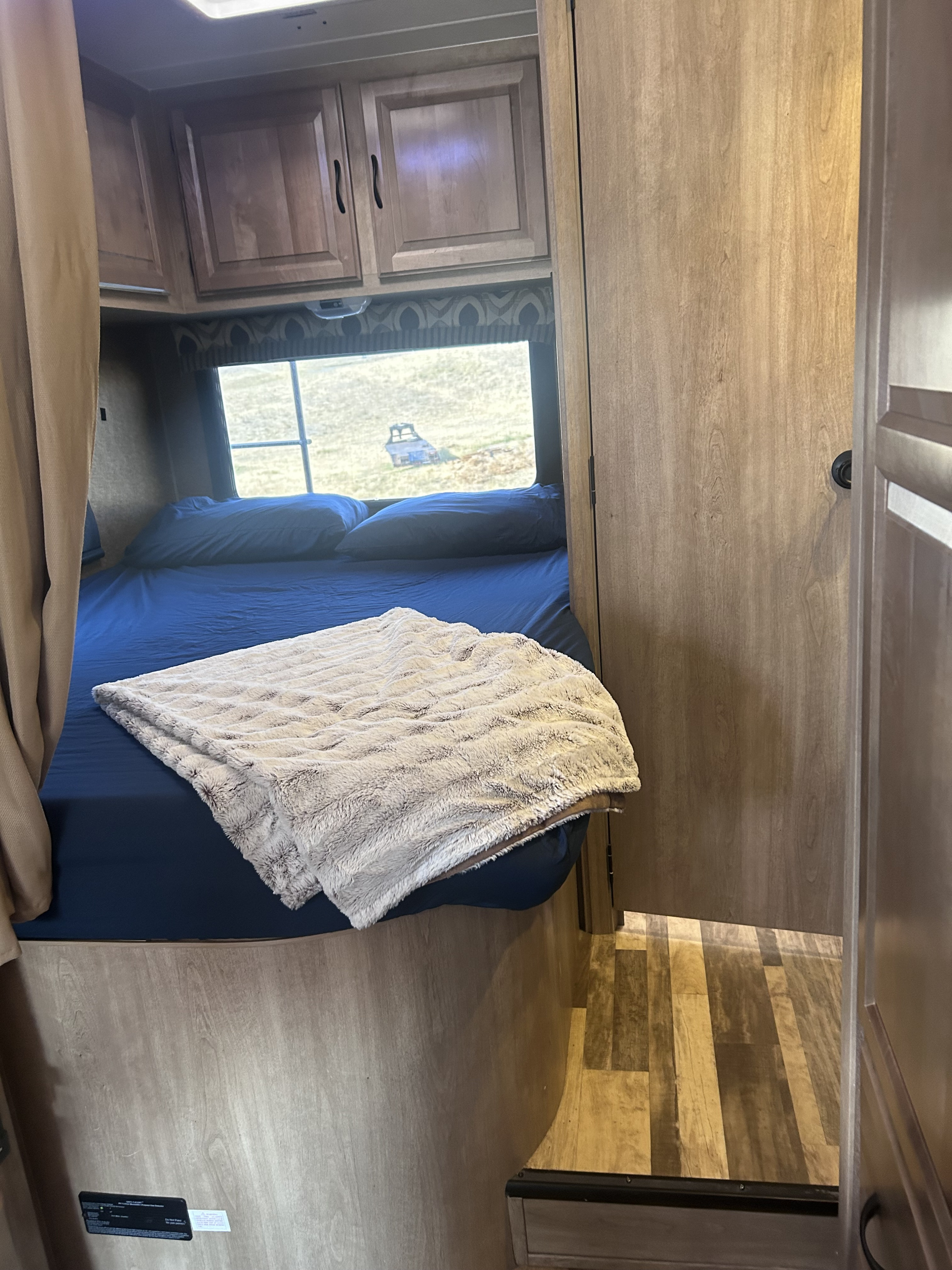Inside an RV rental: a cozy bedroom with blue sheets, fuzzy blanket, wooden cabinets, and a window view of a field.. Forest River Coachmen Prism 2014