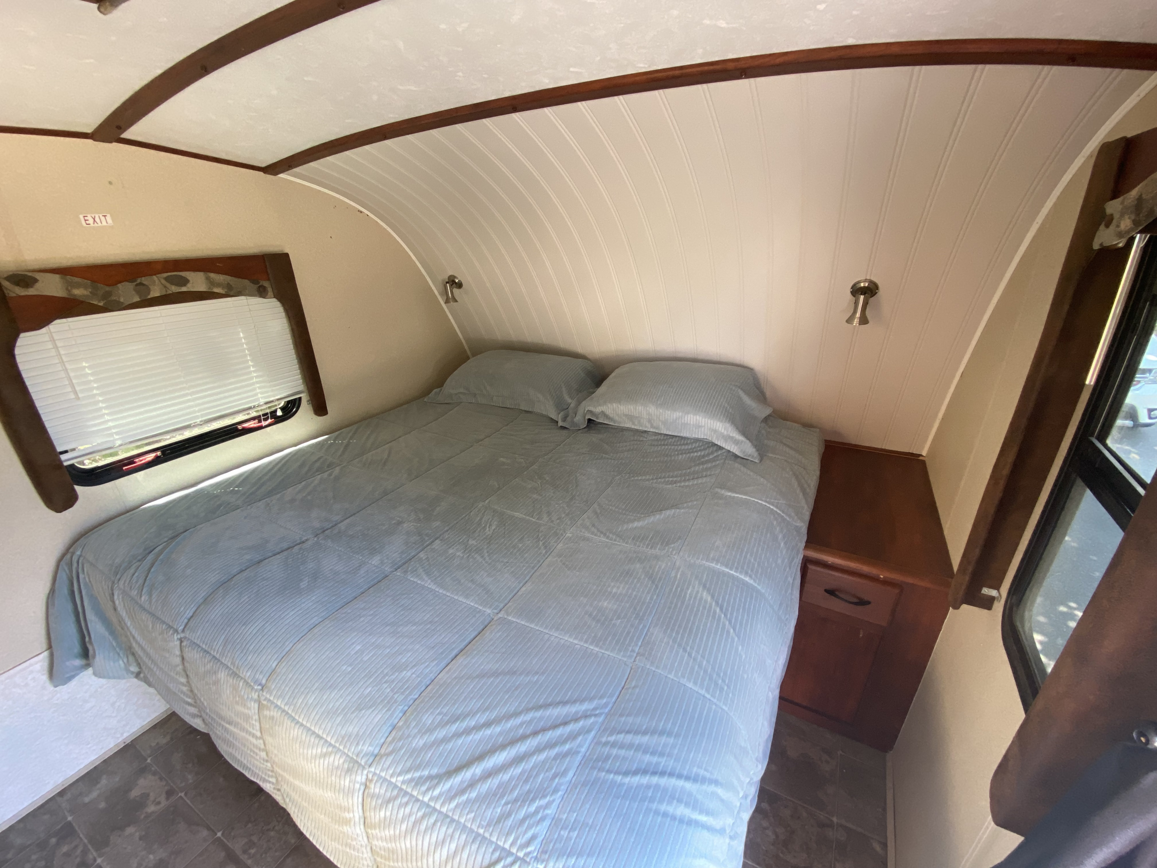 Cozy RV rental bedroom featuring a light blue bed, pillows, a window with blinds, and curved wood paneling ceiling.. MVP Coast 2009
