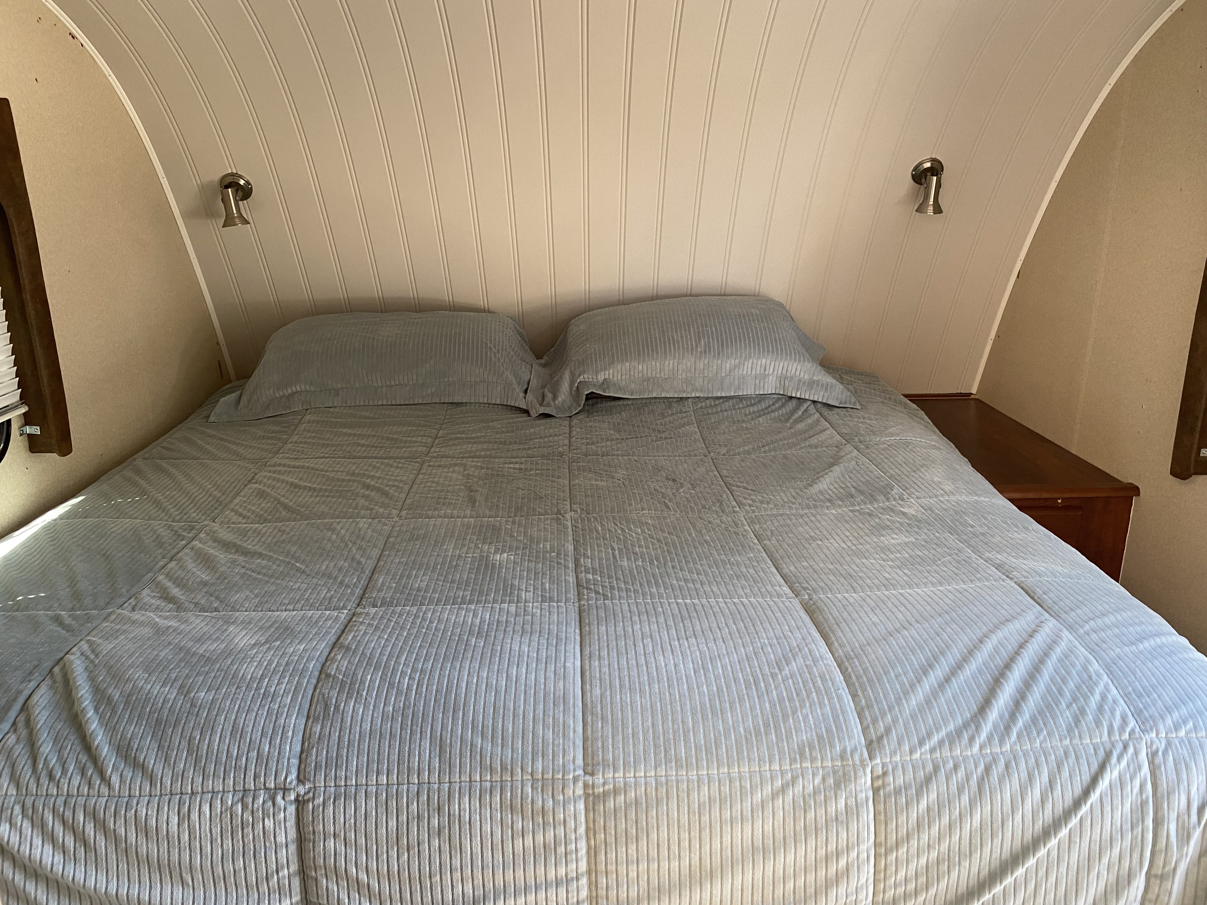 Serene RV rental bedroom with a light grey comforter, pillows, curved paneling, and bedside lights, offering a cozy travel retreat.. MVP Coast 2009