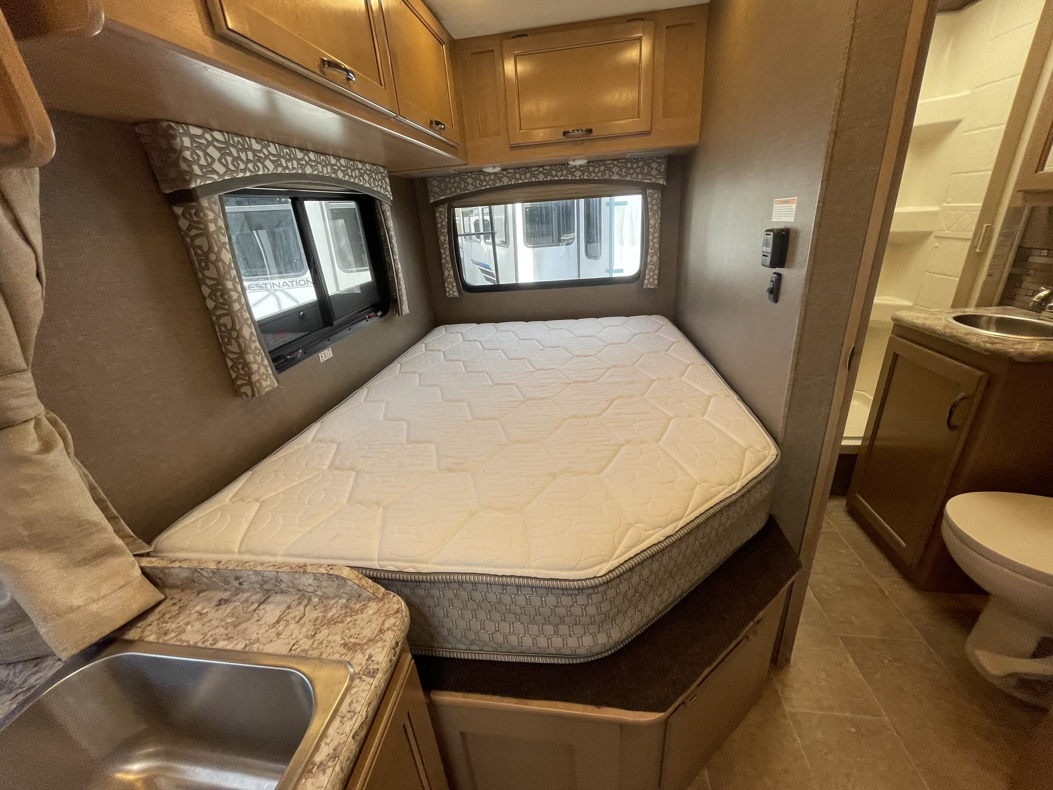 Cozy RV rental bedroom features a bed, two windows, overhead cabinets, and a compact bathroom with toilet and sink.. Thor Chateau 2018