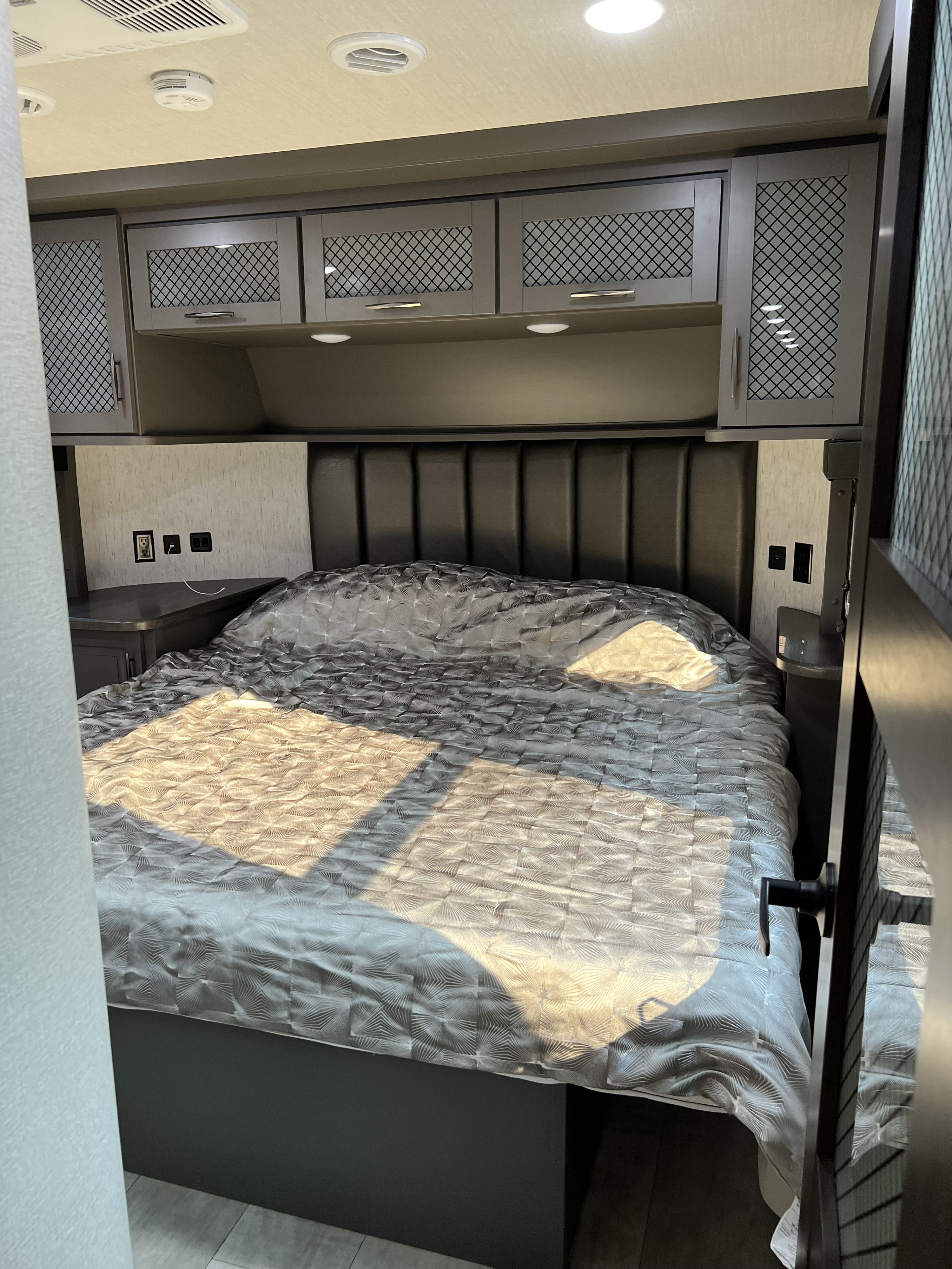 Cozy RV rental bedroom with patterned comforter, padded headboard, and overhead cabinets, illuminated by natural light.. Grand Design Momentum M-Class Toy Hauler 2023