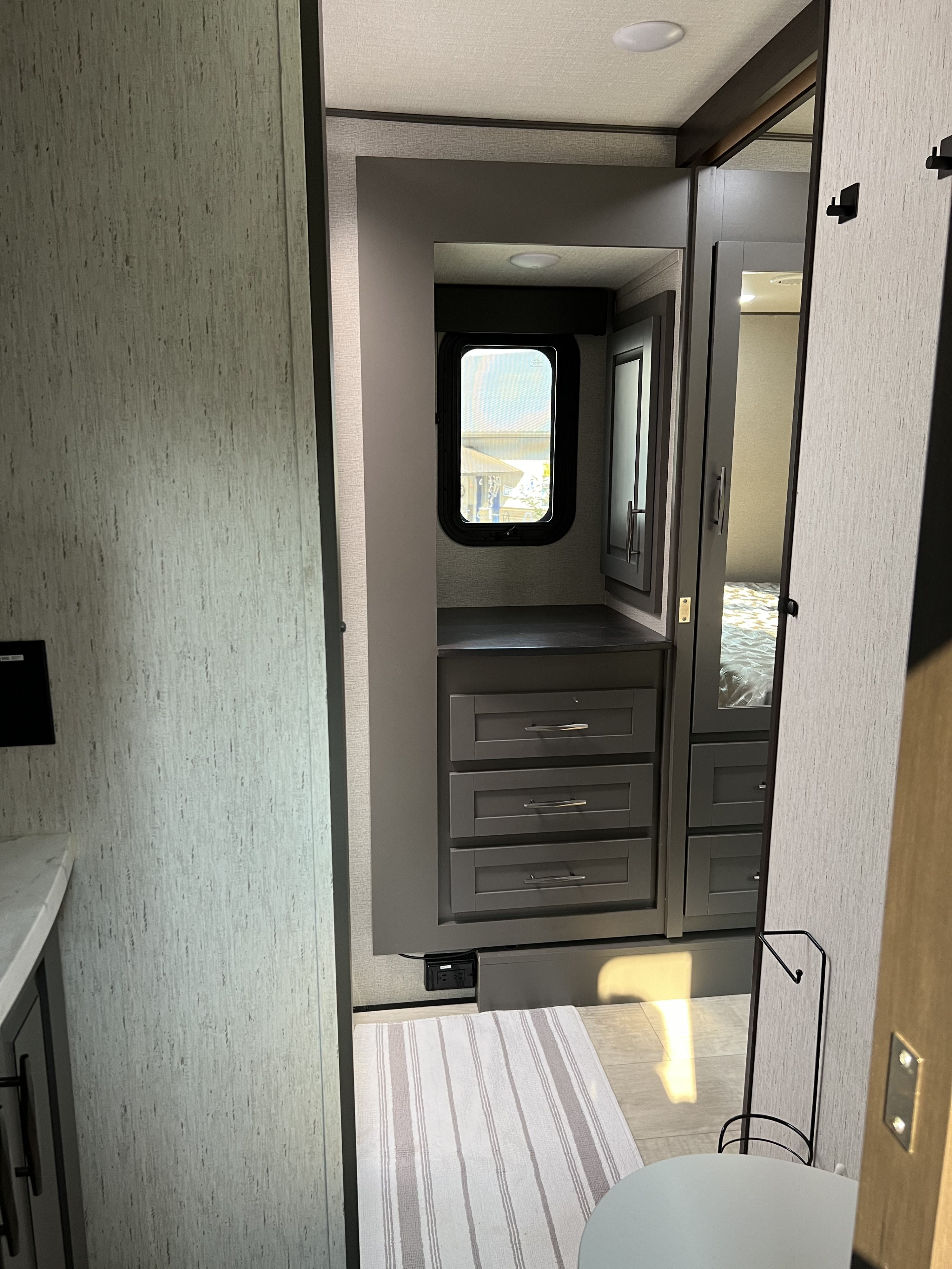 RV rental interior view of a compact dressing area with gray cabinets, window, mirror, and bedroom access.. Grand Design Momentum M-Class Toy Hauler 2023