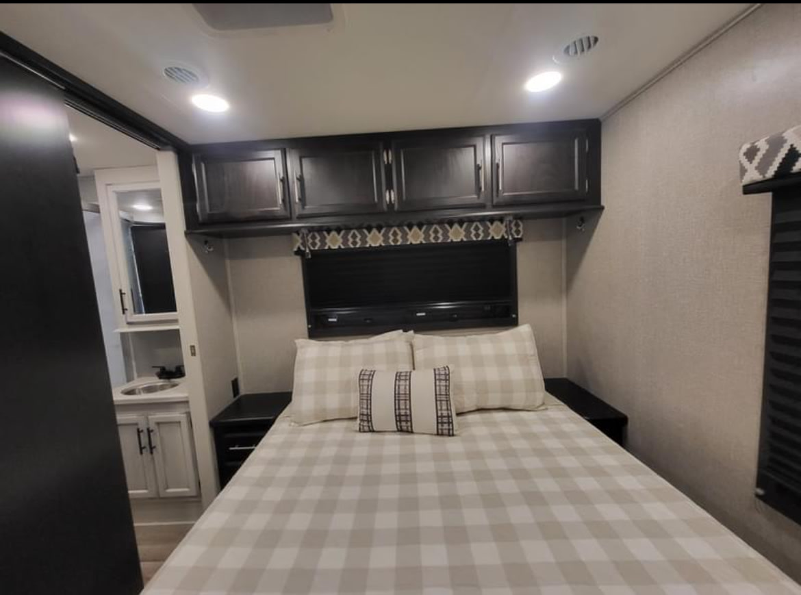 Explore this well-appointed RV rental bedroom, featuring a plaid bed, dark wood storage, and a compact attached bath.. Entegra Coach Odyssey 2021