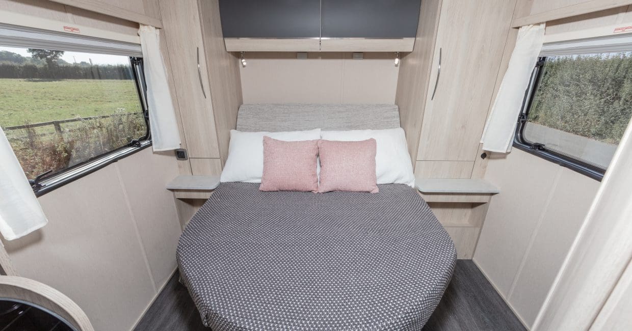 Inside an RV rental: a light-toned bedroom features a bed with white and pink pillows, flanked by windows overlooking a field.. Auto-Trail C71 Expedition 2025
