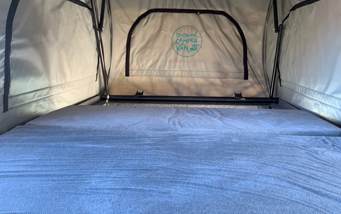 Interior of a pop-top RV rental, featuring a comfortable grey bed and "Colorado Camper Van" logo visible on the back fabric.. Ford Transit Custom 2019