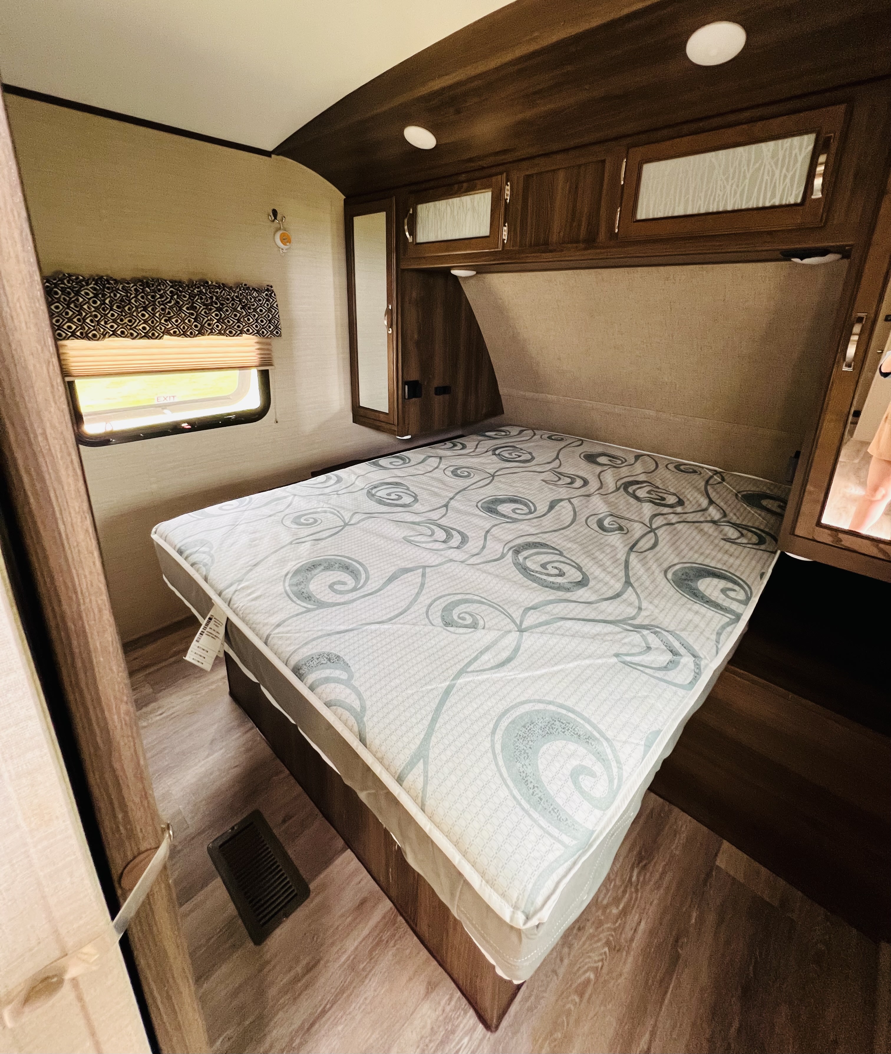 RV rental master bedroom: A compact space with a swirl-patterned mattress, dark wood overhead cabinets, a window, and mirror.. Keystone RV Bullet Ultra Lite 2019