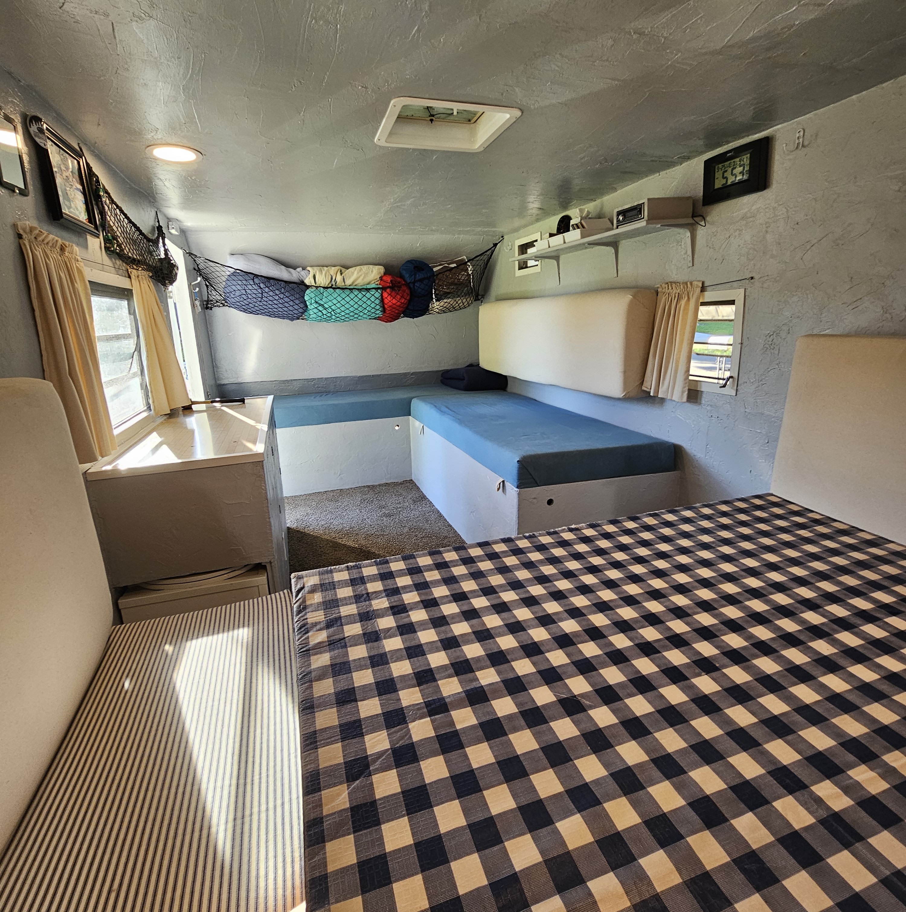 Cozy interior of an RV rental showing benches, sleeping areas with blue cushions, a checkered bedspread, curtains, and overhead storage.. Yellowstone B21539 - 19' 1967