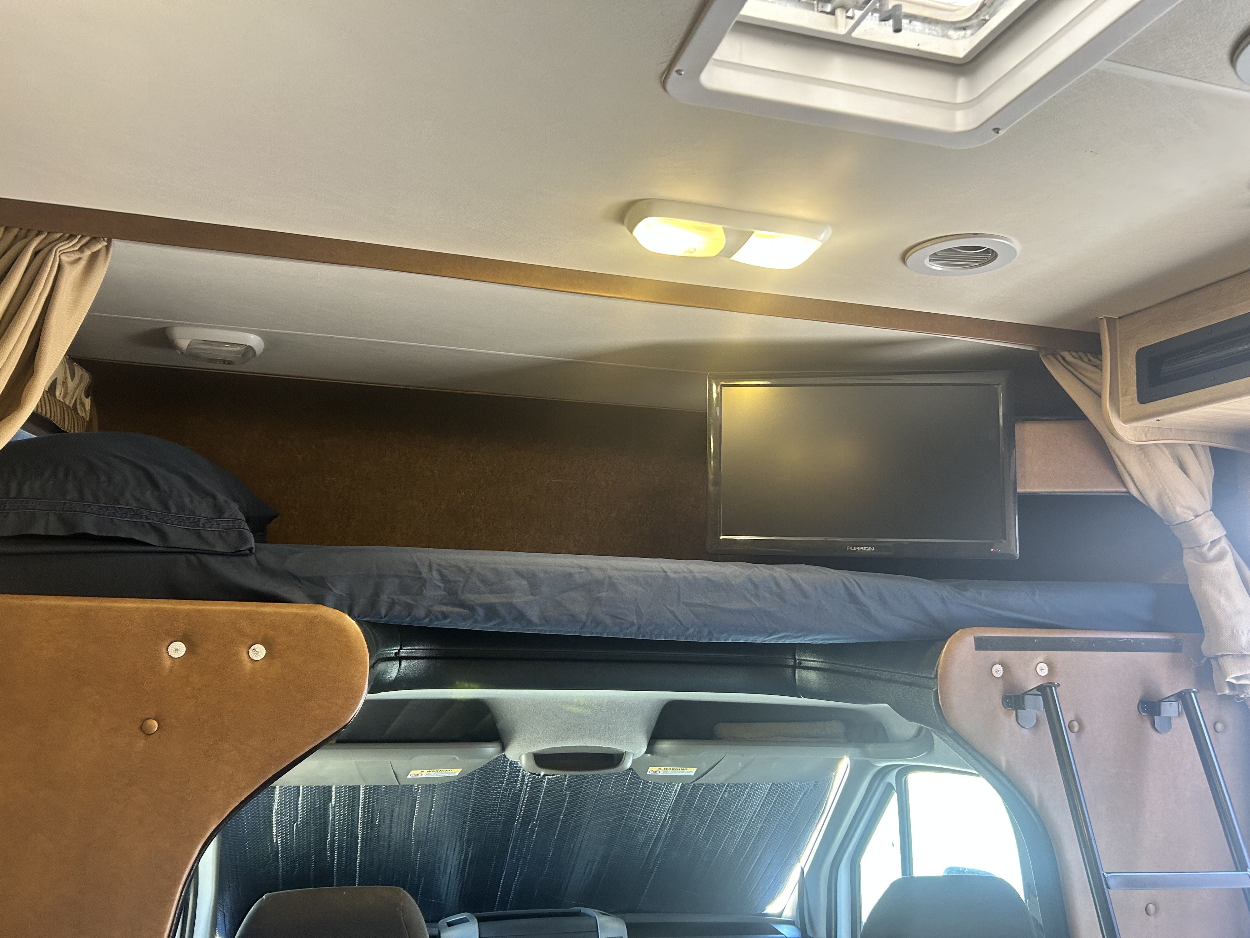 Inside an RV rental: view of the cab-over bunk with dark bedding and a TV, bathed in bright overhead light.. Forest River Coachmen Prism 2014