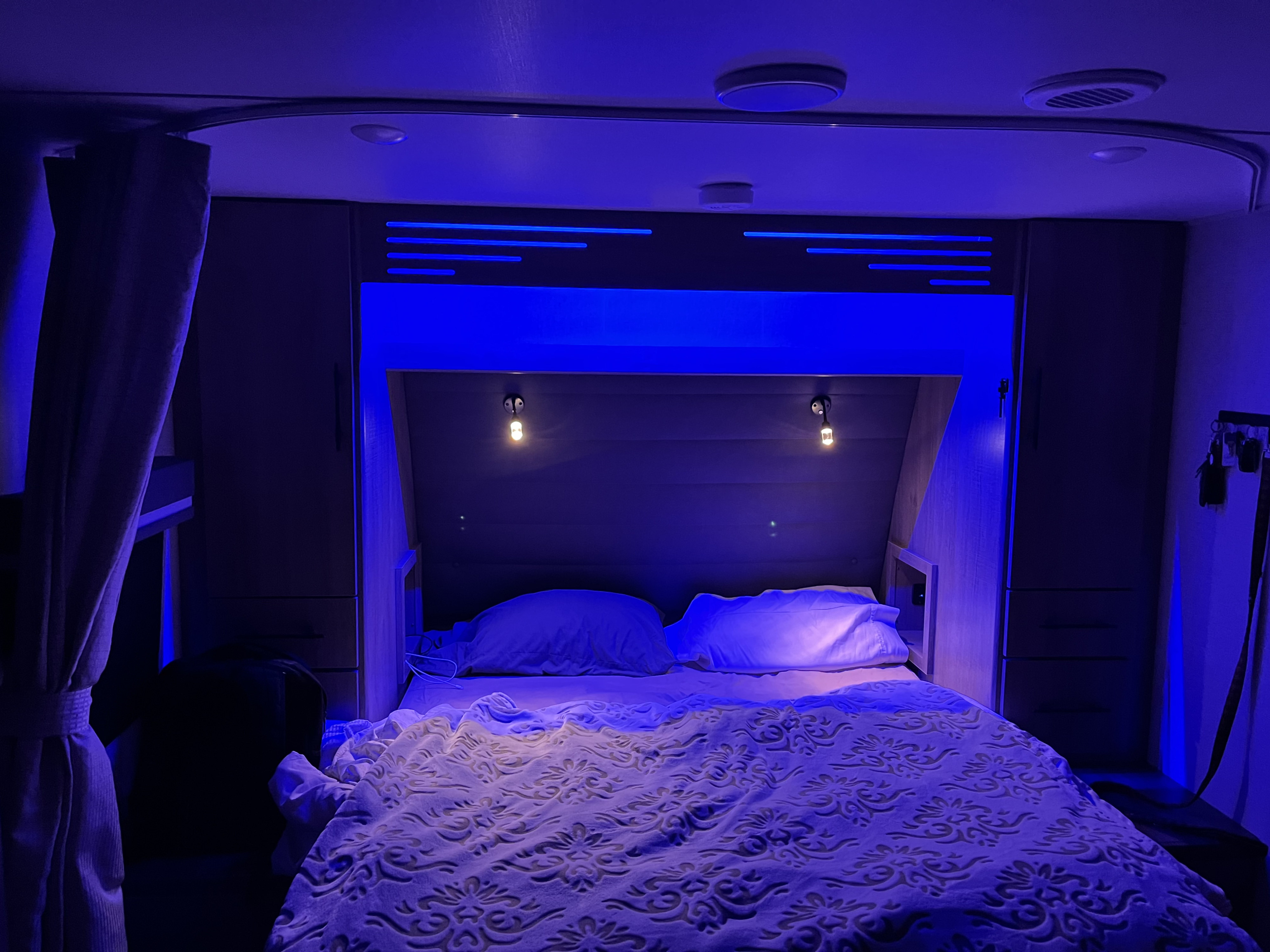 A modern RV rental bedroom illuminated by cool blue LED accents and warm reading lights, showcasing compact living.. Grand Design Imagine XLS 2023