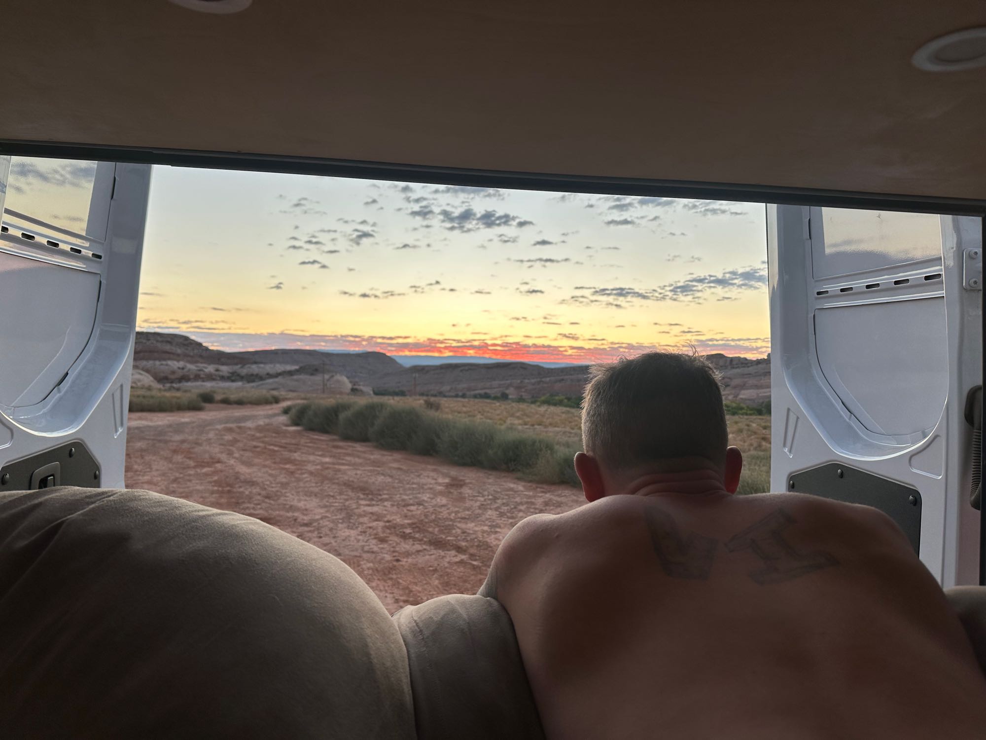 Rear view of a man relaxing inside an RV rental, gazing at a beautiful desert sunset framed by the open doors.. Mercedes-Benz Sprinter 2021