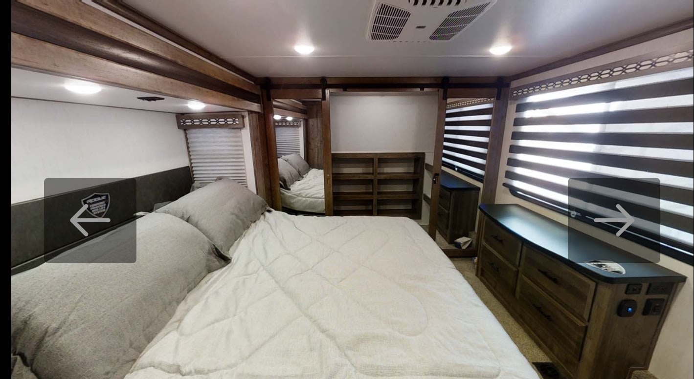 Step into this spacious RV rental bedroom with a comfortable bed, wooden accents, dresser, and bright window blinds.. Forest River Vengeance Rogue Amored Toy Hauler 2021