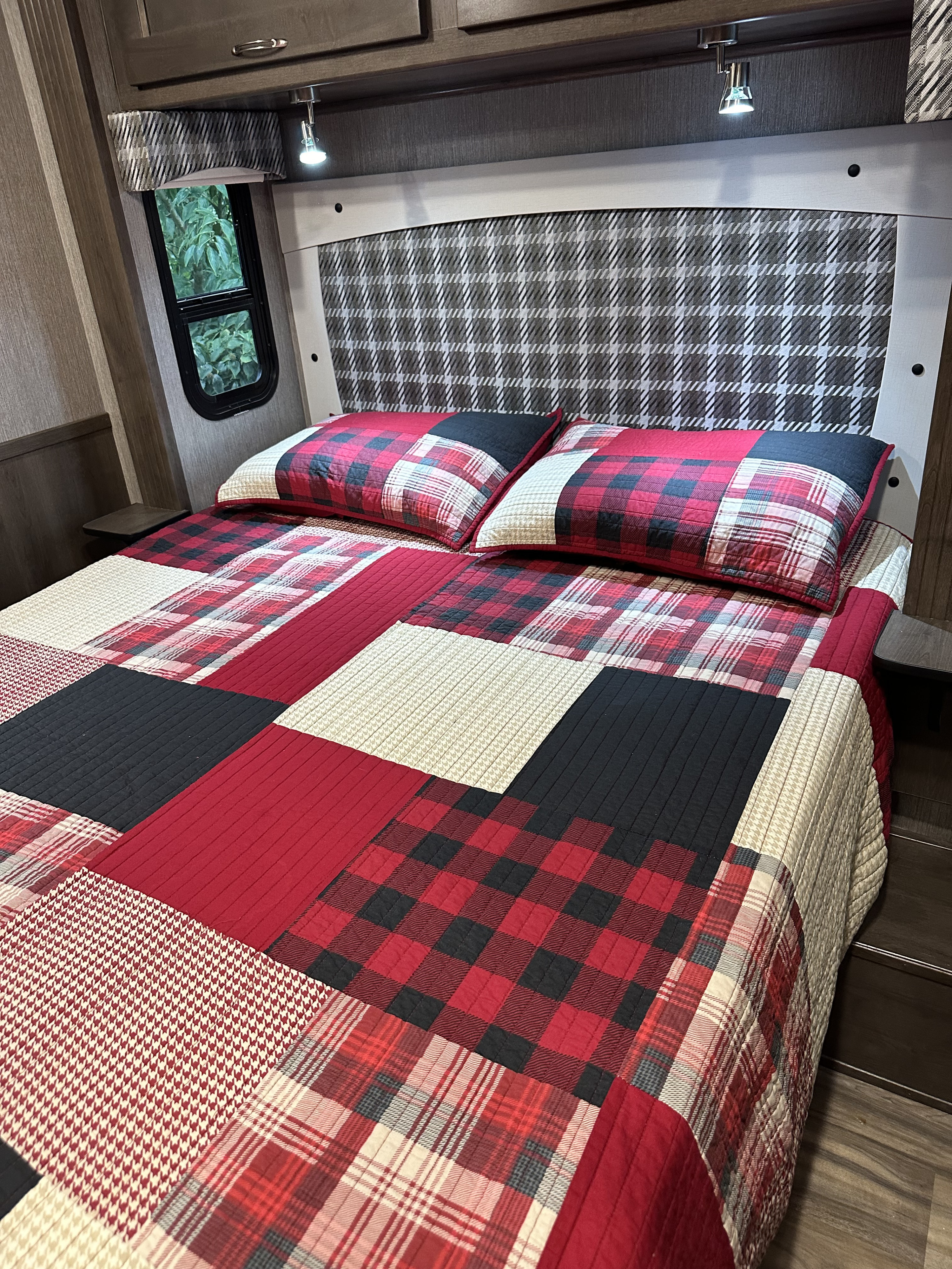 Cozy RV rental bedroom features a vibrant red, black, and white plaid patchwork quilt, matching pillows, and a window view.. Nexus RV Wraith 2022