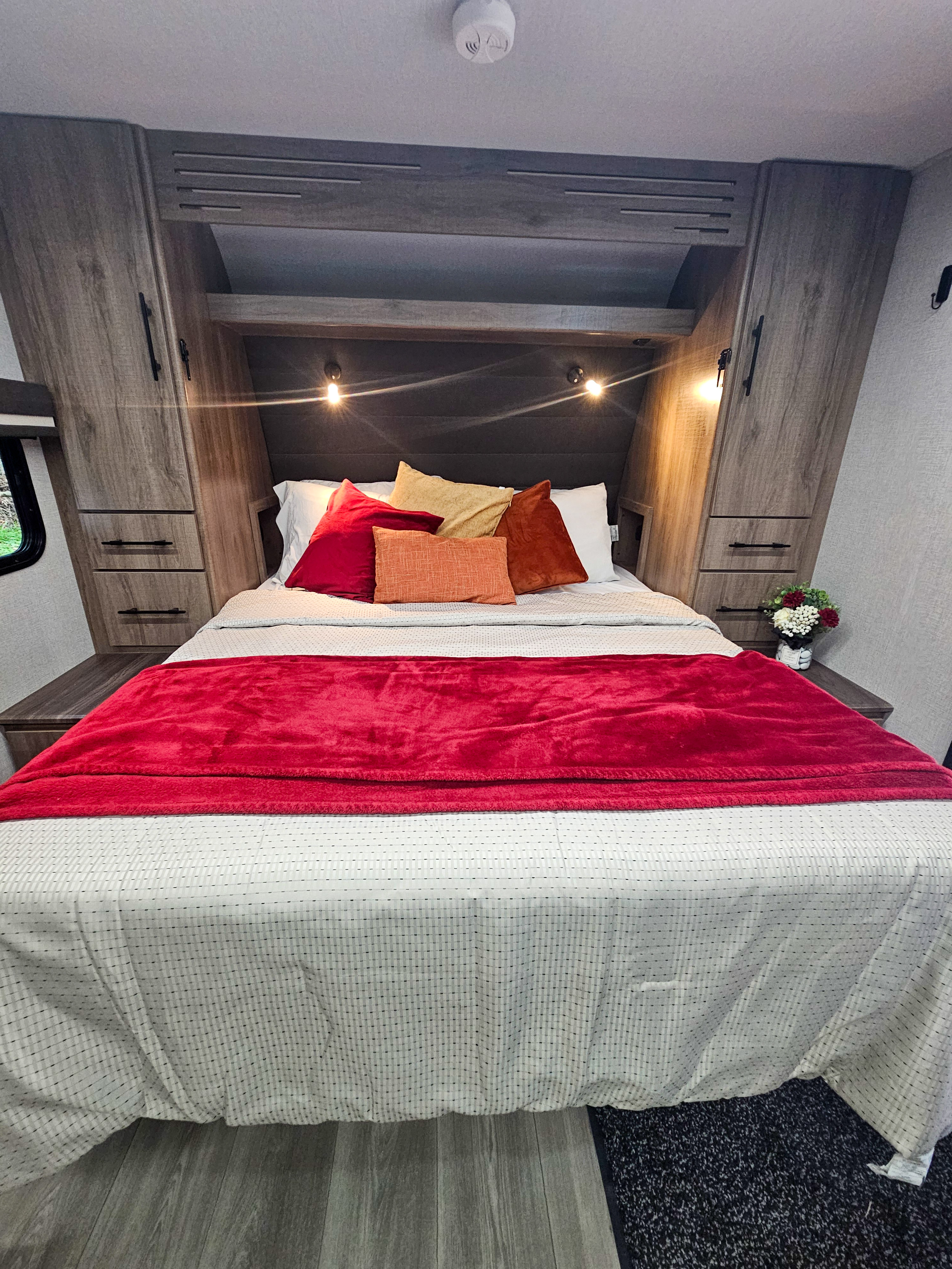 Cozy RV rental bedroom featuring a full bed with a red blanket, built-in cabinets, and warm wall lighting.. Grand Design Imagine XLS 2024