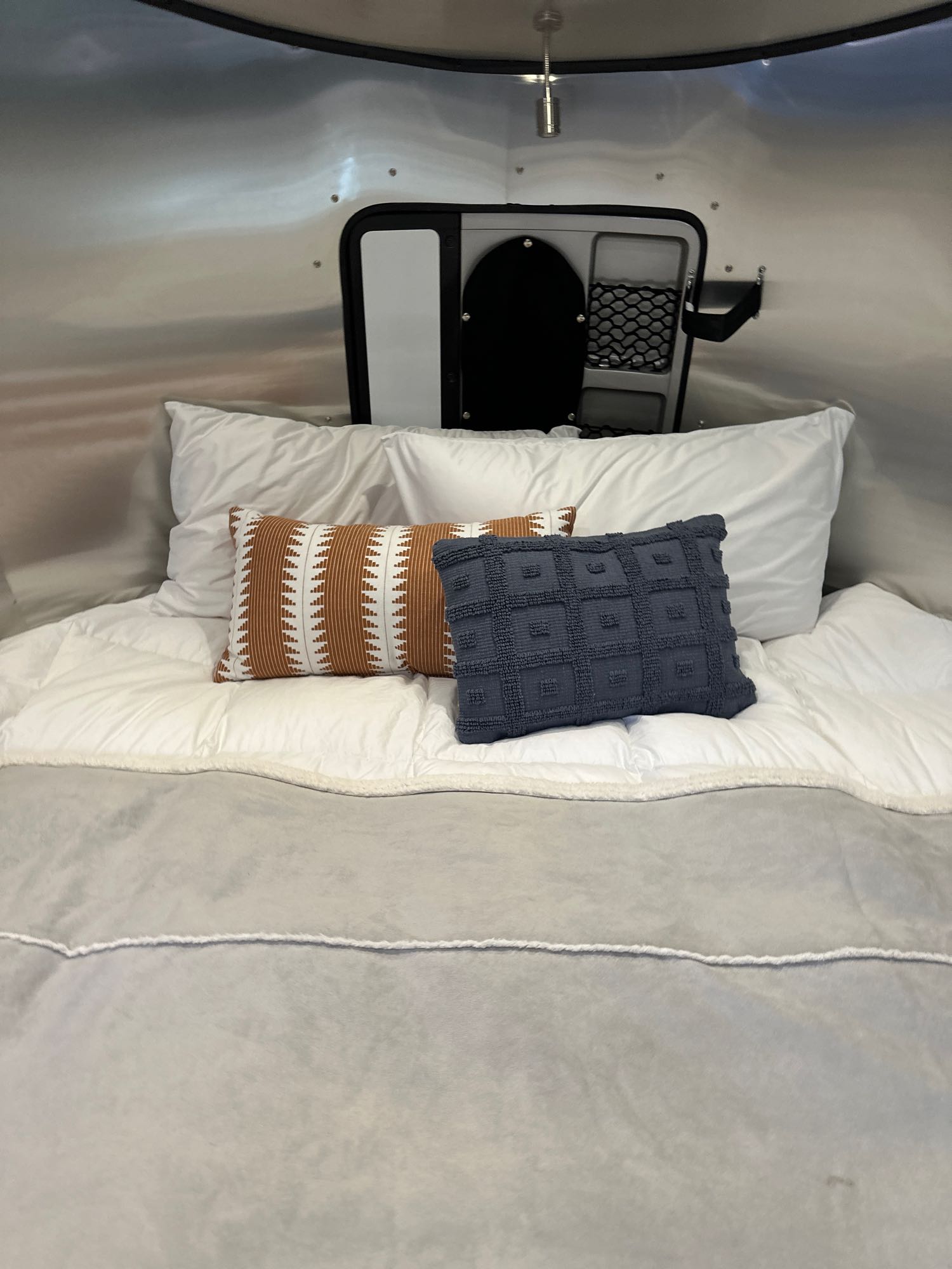 Plush bed in an RV rental, featuring white bedding, an orange geometric pillow, and a blue textured pillow.. Airstream Basecamp 2023