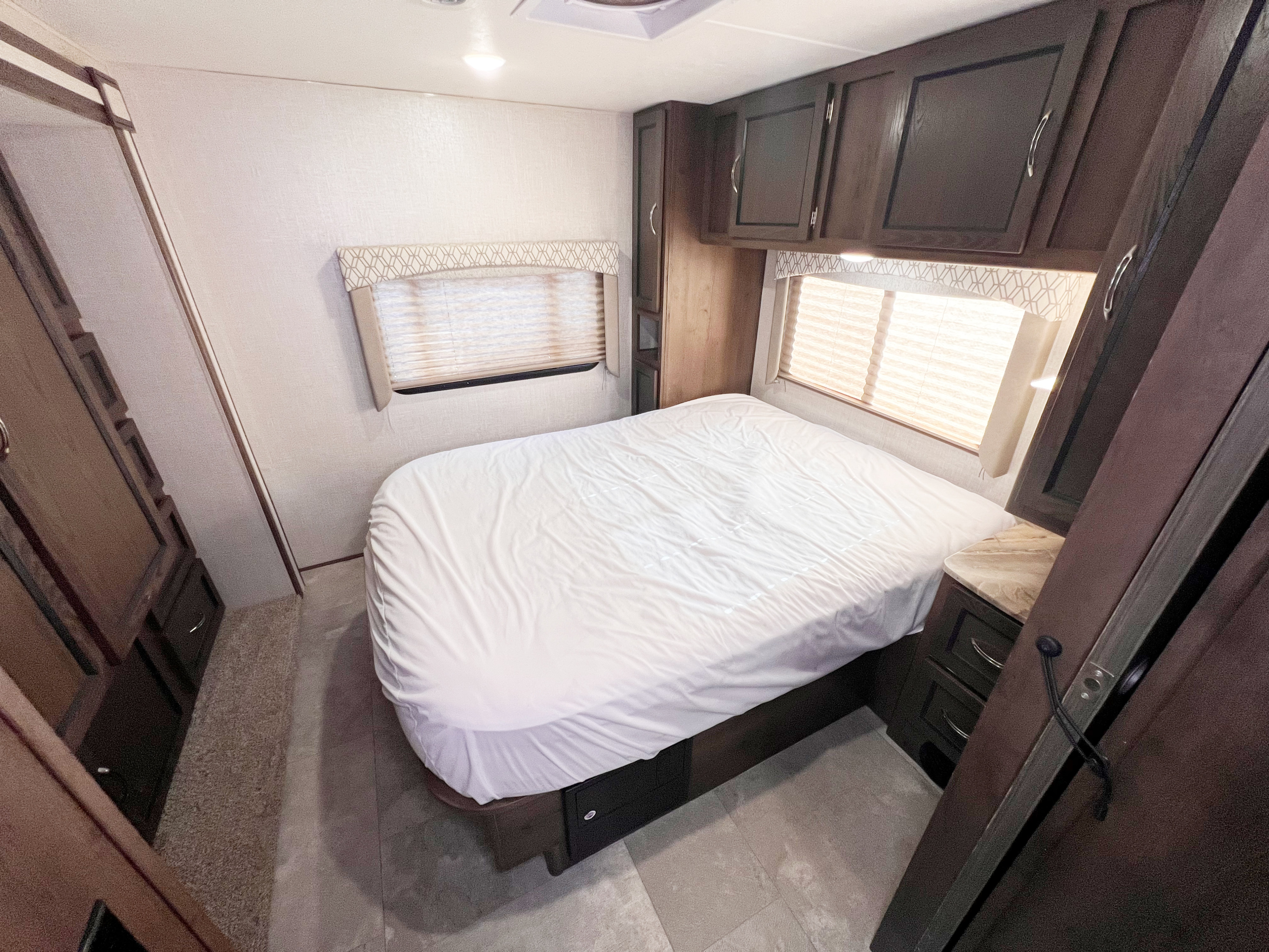 Inside an RV rental: a cozy bedroom with a neatly made bed, dark wood cabinets, and two large windows with blinds.. Forest River Freelander 2020
