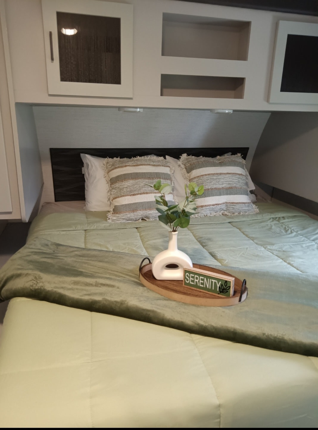 Relax in this RV rental bedroom featuring a comfy bed with green bedding, decorative pillows, overhead cabinets, and a serenity tray.. Heartland RVs North Trail 2022